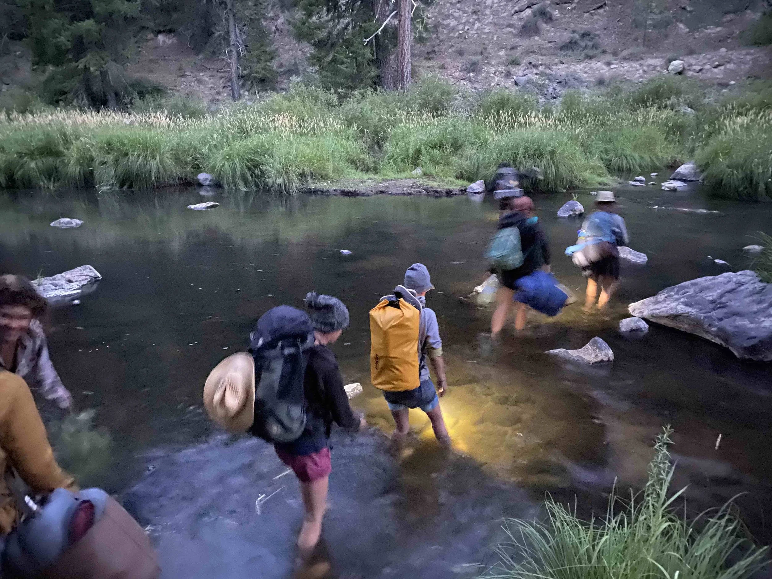 A group of people crossing a shallow river in a forested area at dusk, with trees and grass visible along the riverbanks.