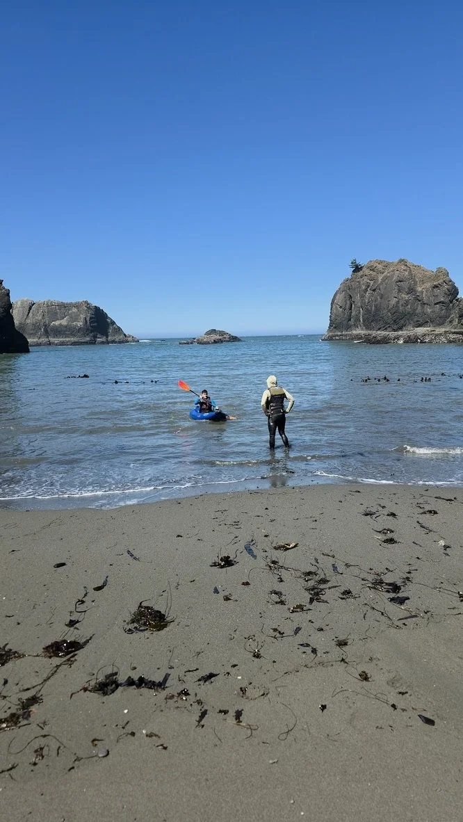 A person in a wetsuit standing at the shoreline with a kayaker paddling in the water nearby, and large rock formations in the distance under a clear blue sky.