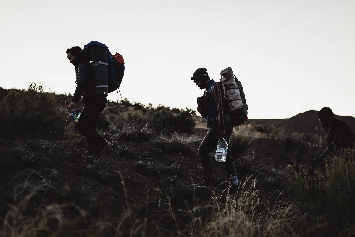 Two hikers with backpacks and water bottles hiking on a mountain trail at dusk or dawn.