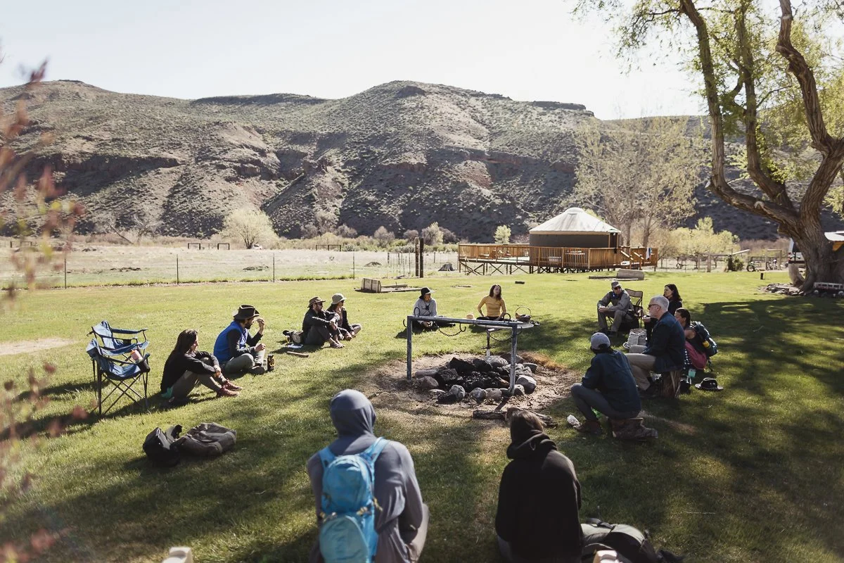Group of people sitting in a circle around a campfire pit on a grassy field, with mountains in the background and a large tree on the right.