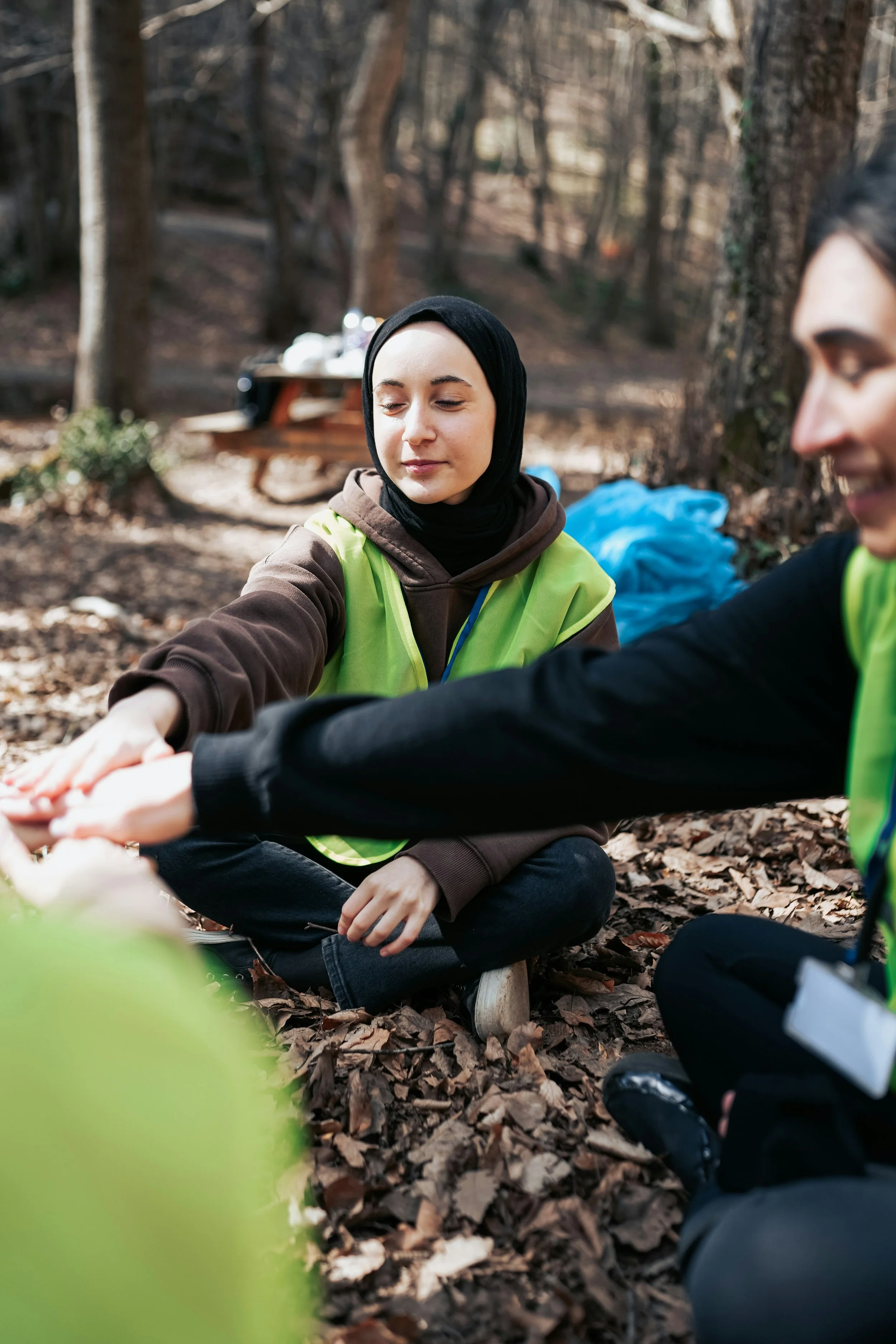 Two women sitting on the ground in a wooded area, wearing yellow safety vests and black hoodies, holding hands.