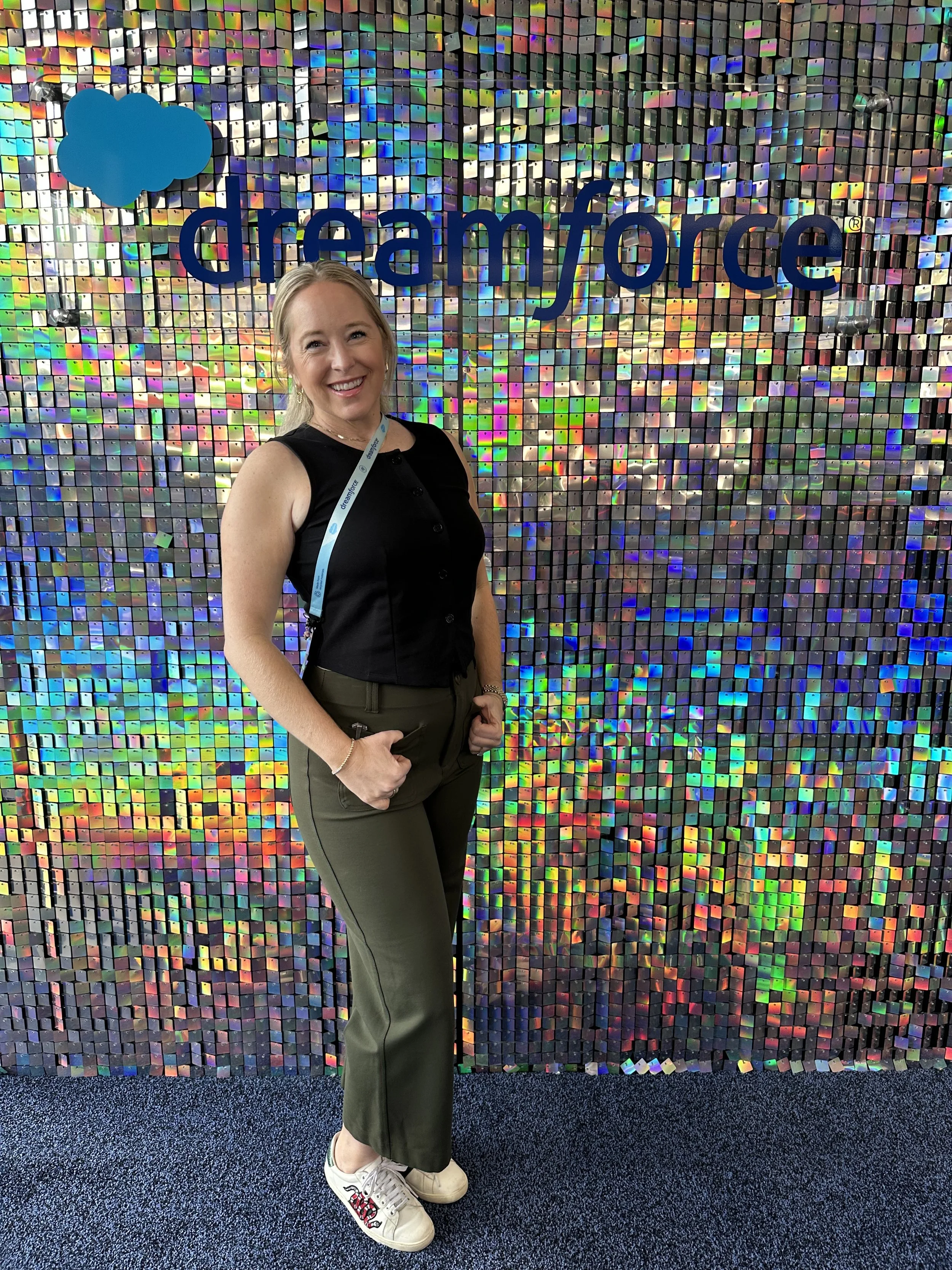 A woman standing in front of a colorful, holographic mosaic wall at Dreamforce event, smiling at the camera.