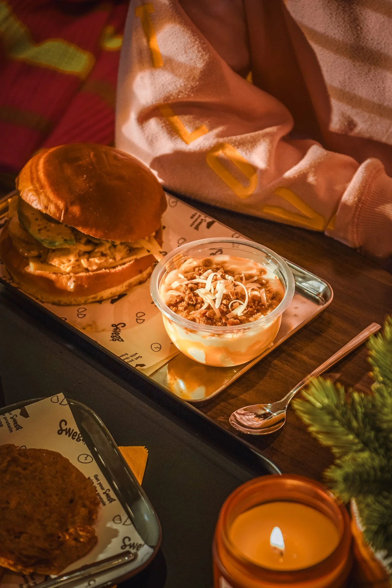 Een tray met een Swegg Sandwich, een schaaltje met pudding en een lepel, een kaars in een pot en een beetje groen decoratie op een houten tafel.