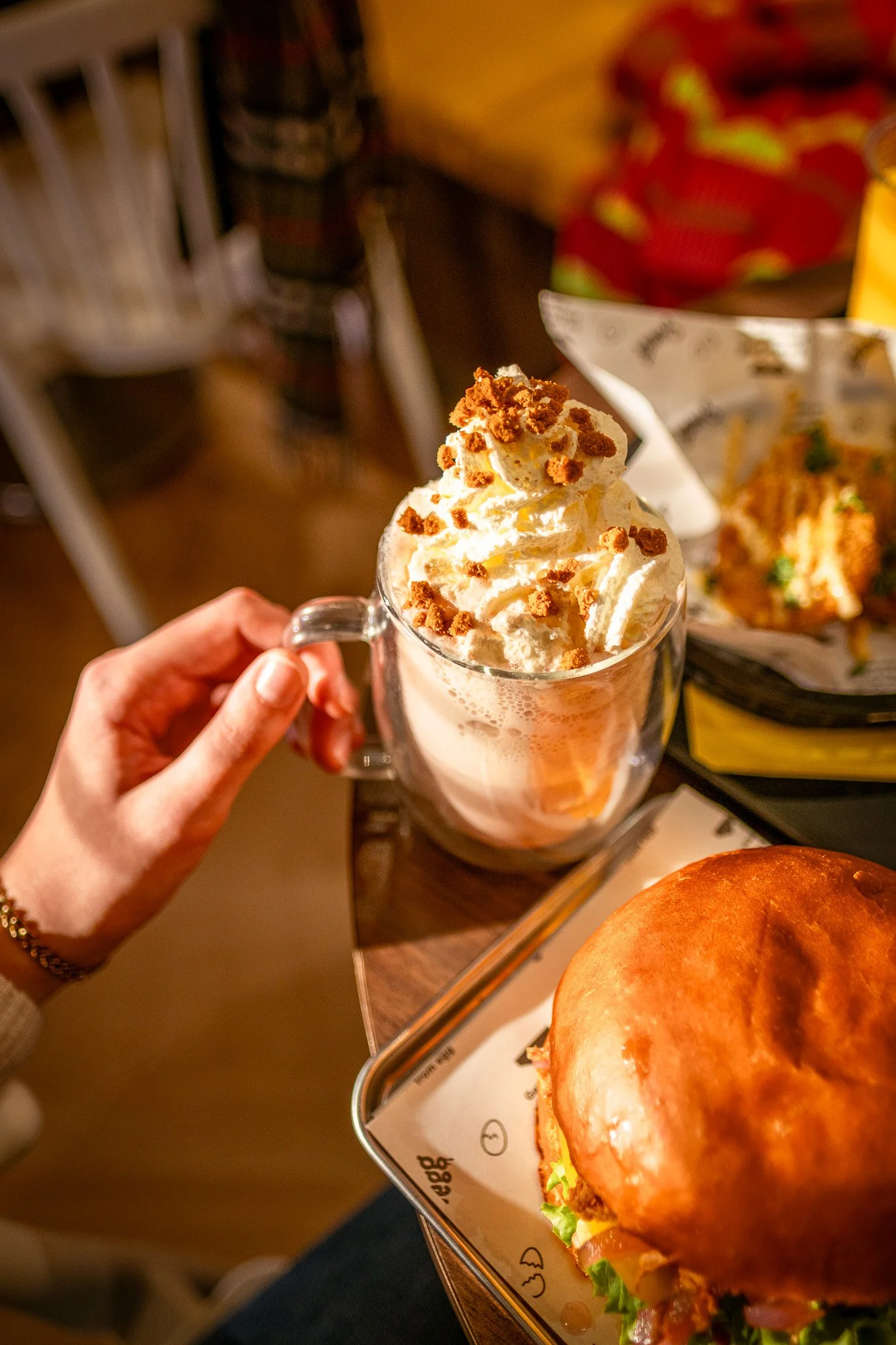 Een hand houdt een glas met een warme chocolademelk met slagroom en kruimels, op de achtergrond een Swegg Sandwich en een bak met friet.