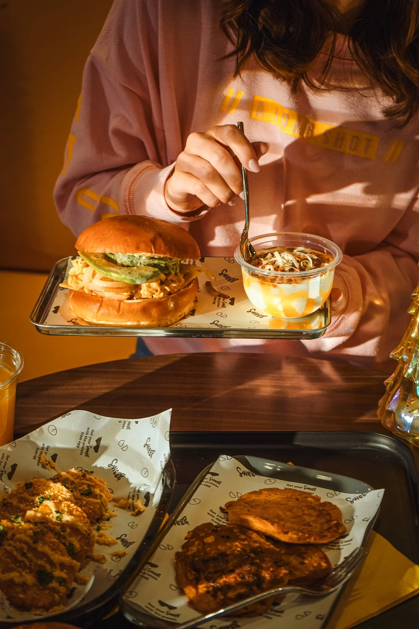 Person die een burger en een gerecht met saus eet, op een dienblad, met ander eten op de tafel zoals aardappelballetjes en vlees bovenop, in een restaurant.