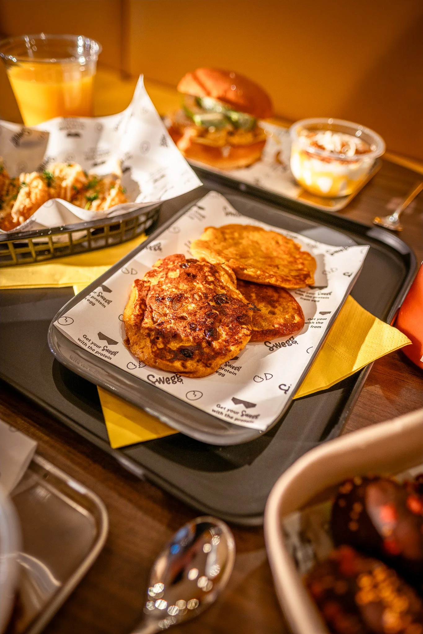 Ontbijtschotel met gebakken ei en aardappelkoek, daarnaast een hamburger met groenten en saus, en een bakje met lauwwarme yoghurt en fruit.