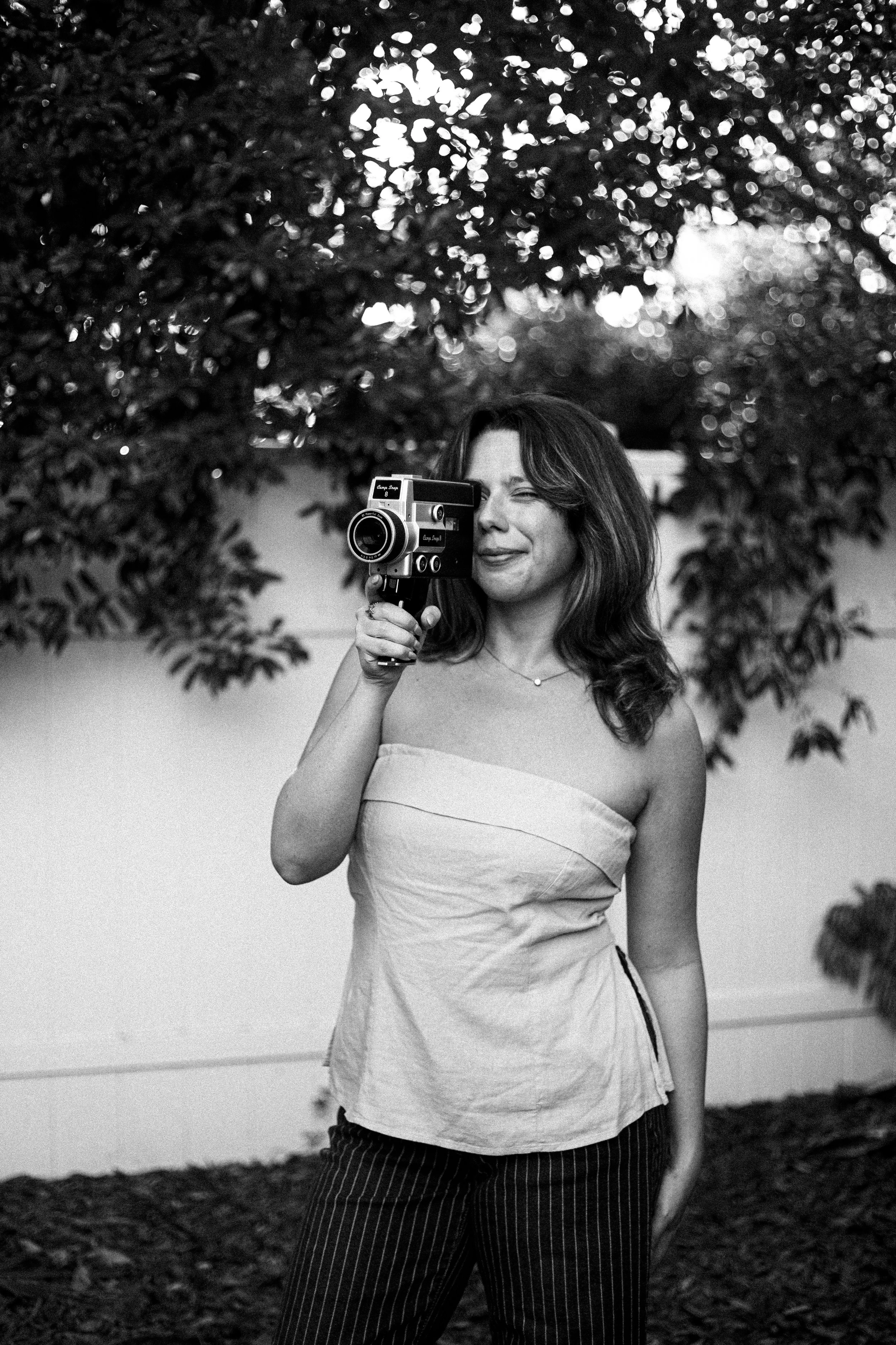 A woman holding a vintage camera and smiling outdoors in black and white, with trees and leaves in the background.