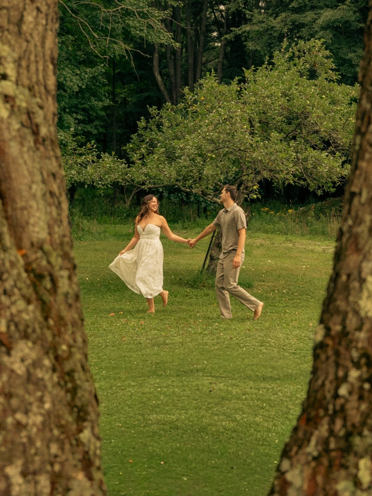 A couple dancing and holding hands in a lush green outdoor setting, viewed through two tree trunks in the foreground, with the woman in a white dress and the man in casual attire, under a tree.
