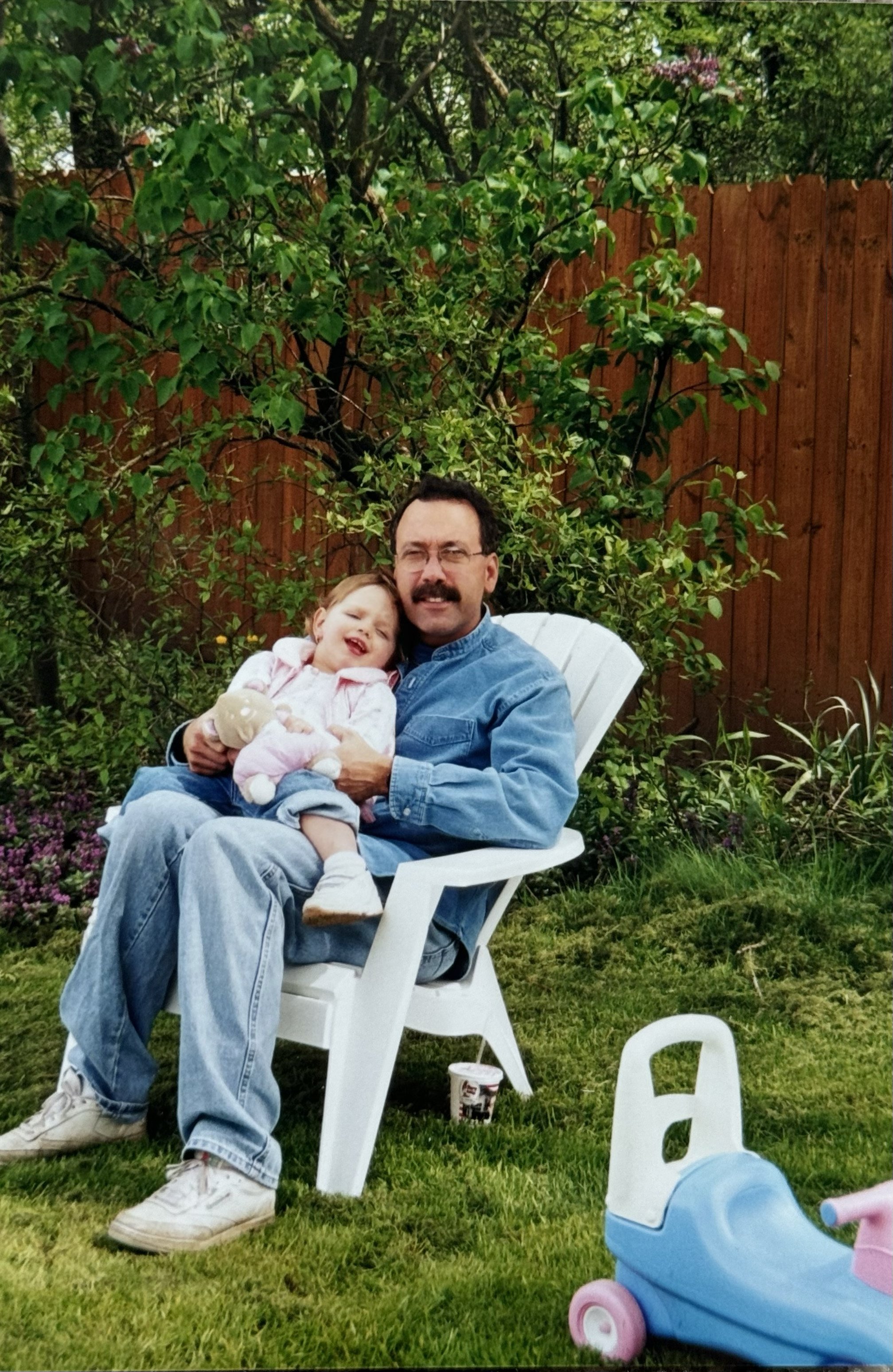 A man with glasses and a mustache holding a young girl on his lap, both smiling and sitting outdoors in a garden with green plants and a wooden fence. The girl appears to be laughing and playing with a stuffed toy. There is a white plastic chair and a pink and blue toy stroller on the grass.