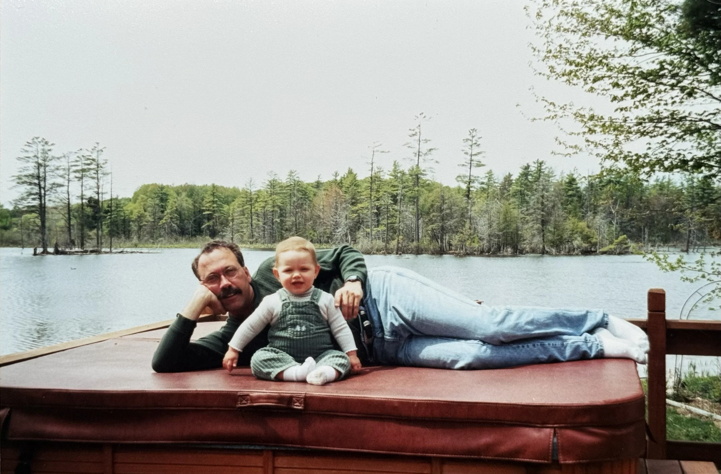 A man with a mustache and glasses lying on his side on a wooden dock by a lake, with a young child sitting on his lap, both facing the camera. The background shows a lake with trees and a cloudy sky.