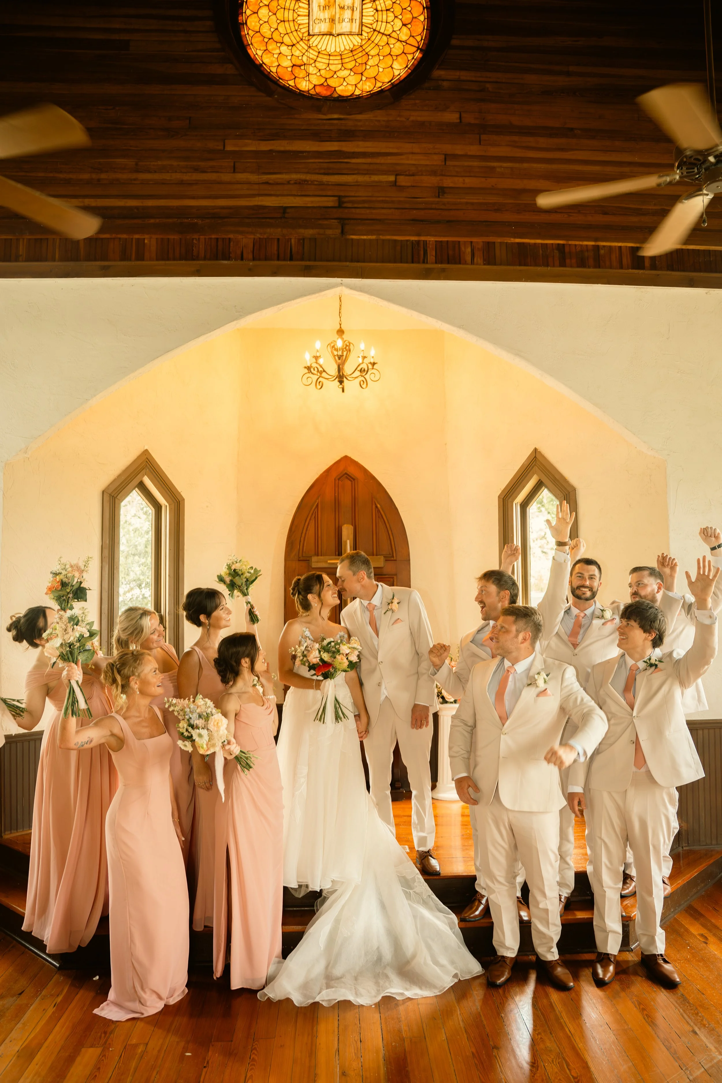 A wedding ceremony inside a church in Dunedin, FL with a bride and groom at the altar surrounded by bridesmaids and groomsmen, some raising their hands in celebration.
