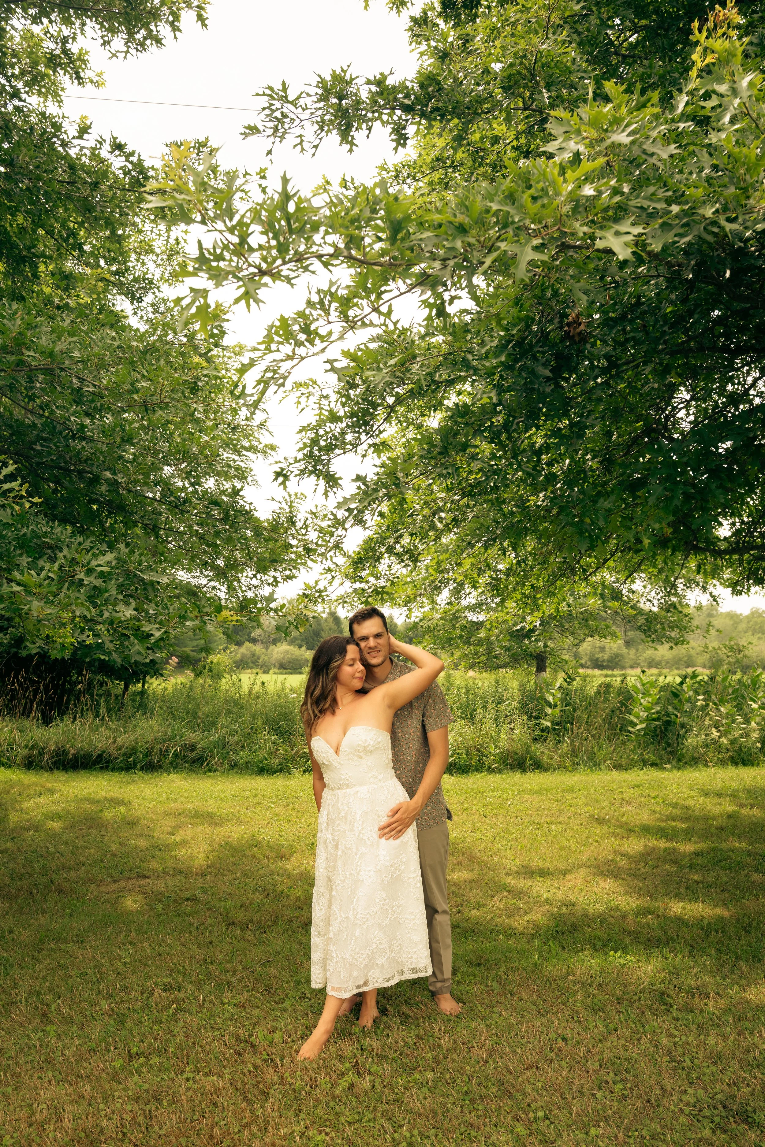 A bride and groom standing on grass in a park, embracing under a large tree with lush green leaves, during daylight.