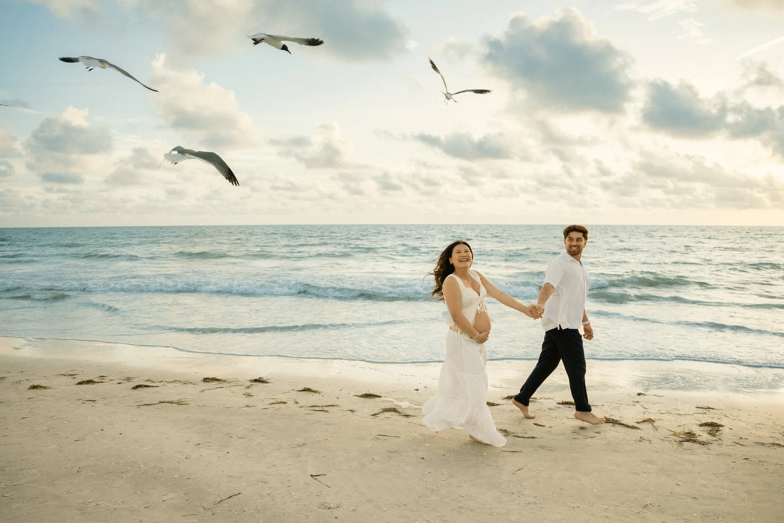 A couple holding hands and walking along Clearwater Beach with seagulls flying overhead and the ocean in the background