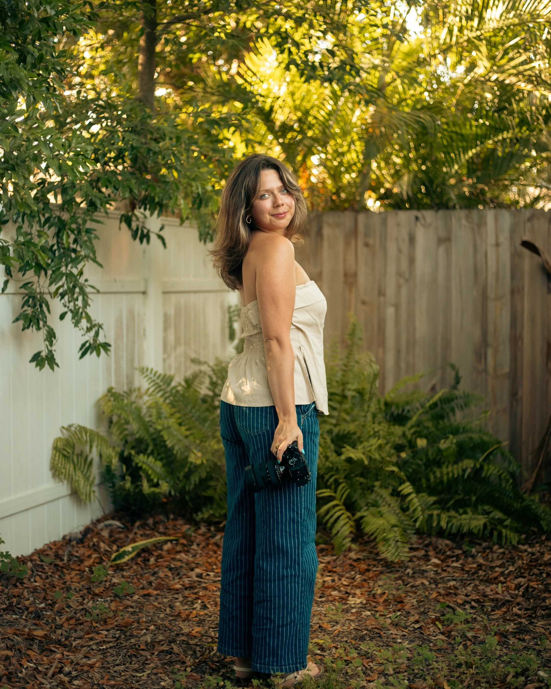 A woman with shoulder-length brown hair standing outdoors on a leaf-covered ground, holding a black camera in her right hand, wearing a strapless beige top and blue striped wide-leg pants, with a wooden fence, green plants, and trees in the background, illuminated by sunlight.