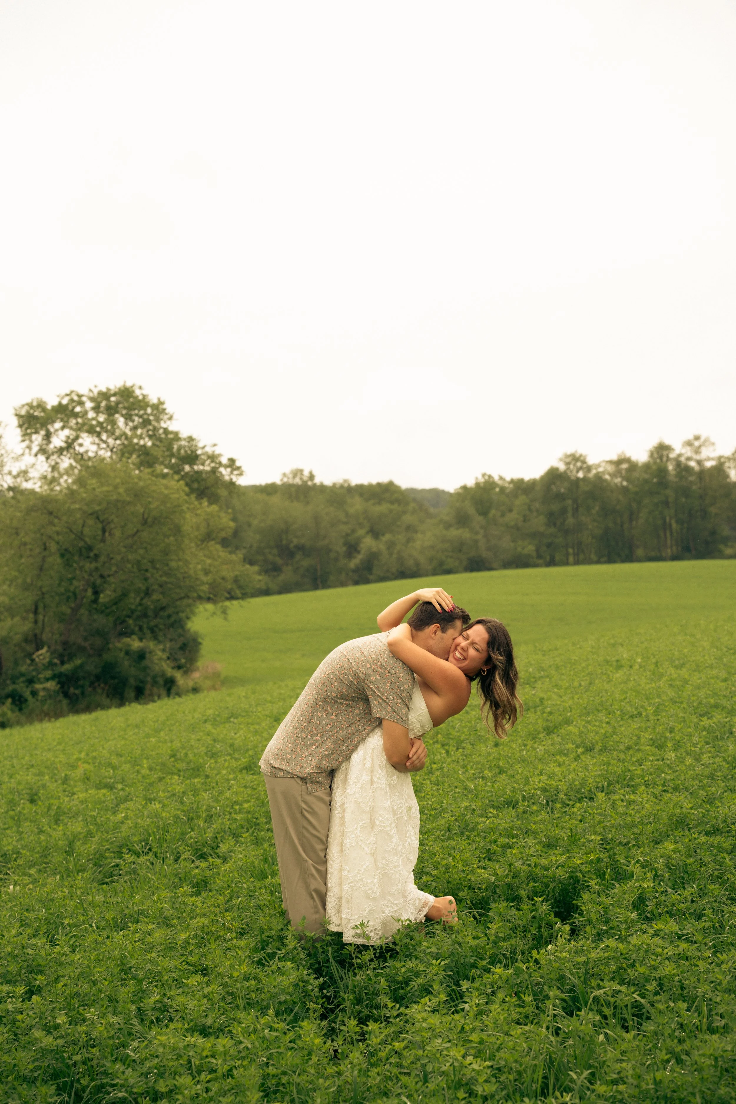 A couple in wedding attire sharing a kiss in a green field with trees in the background under a cloudy sky.