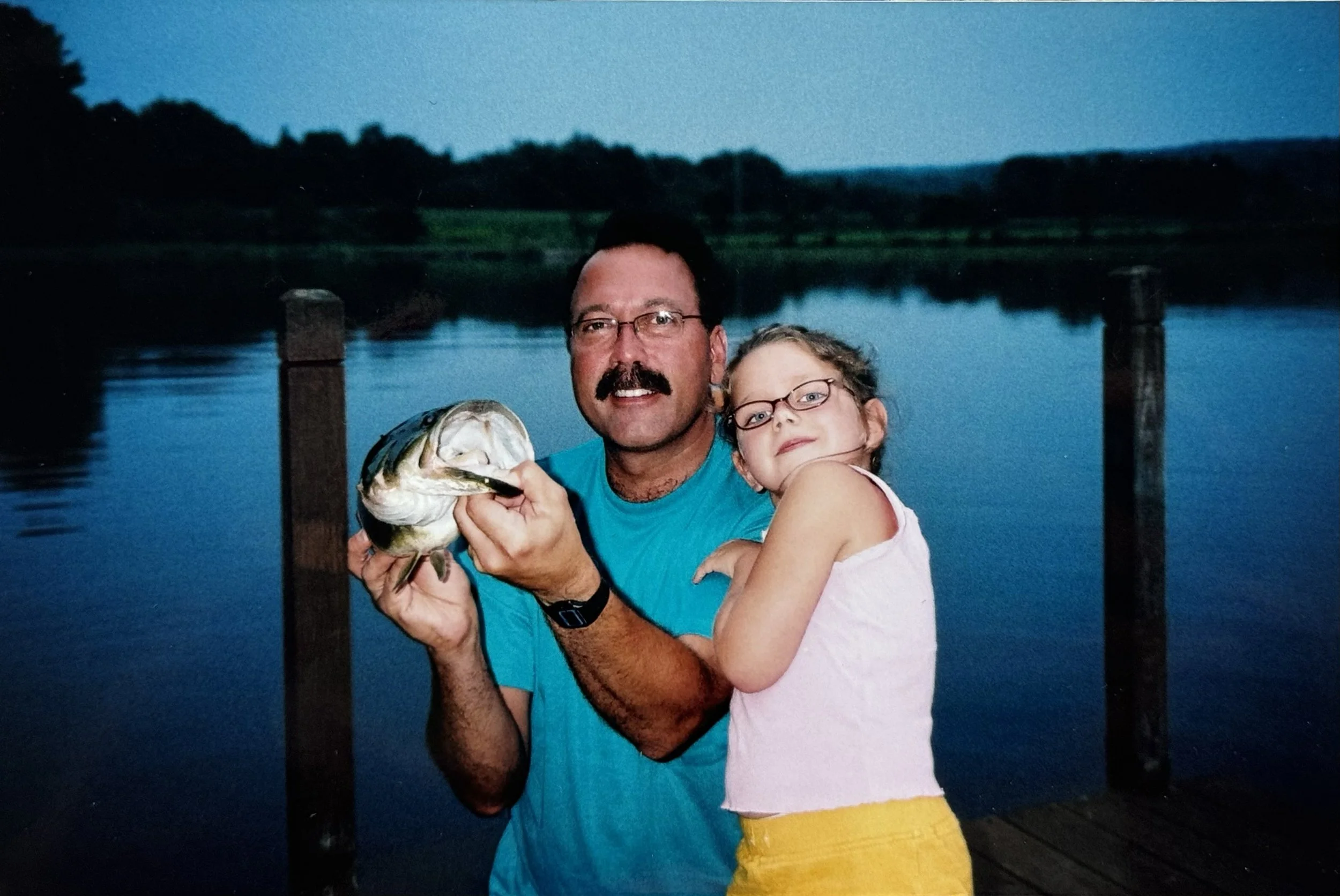 A man with glasses and a mustache holding a large fish, standing next to a young girl on a dock overlooking a body of water during dusk or evening.