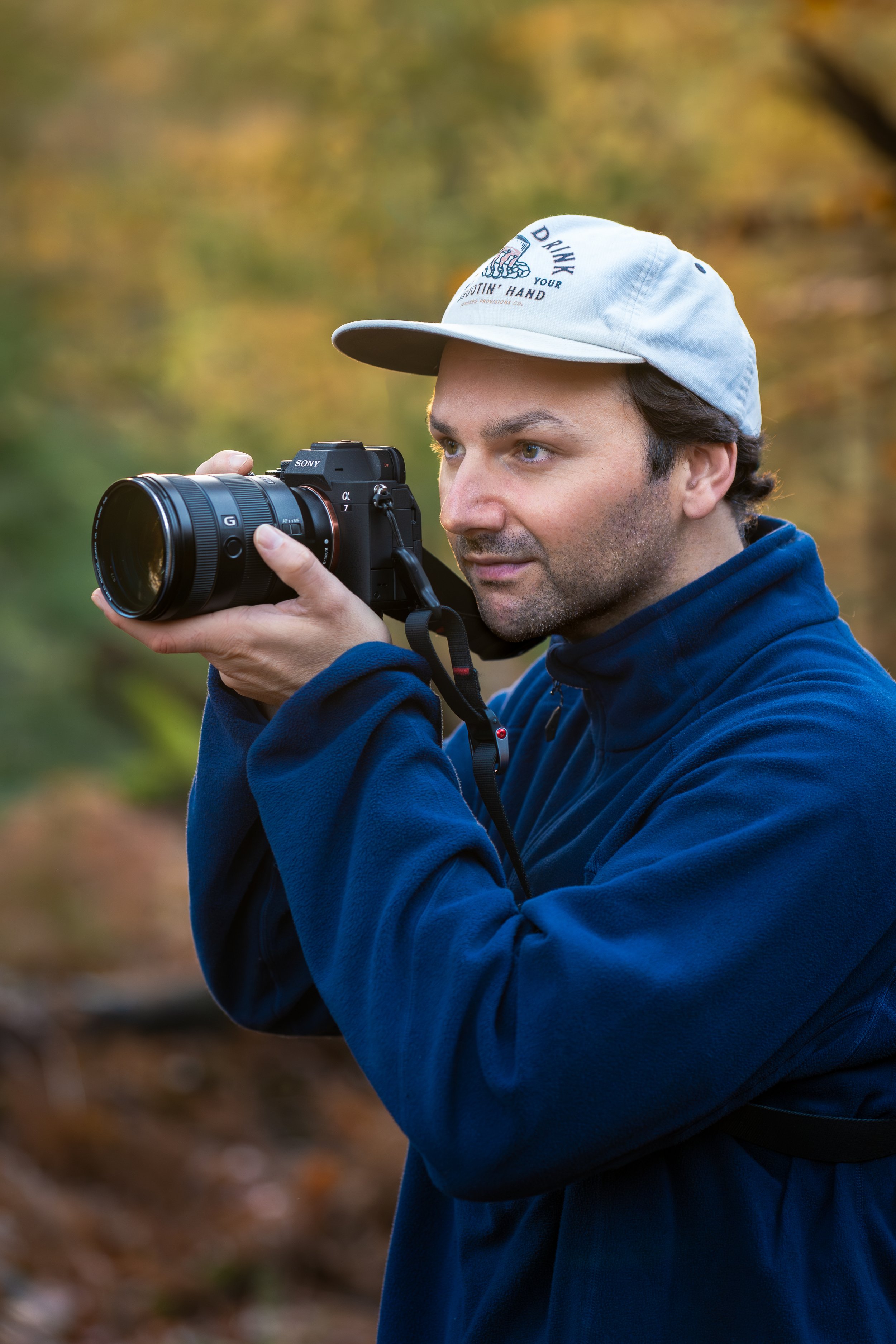 A man in a blue jacket and gray cap is taking a photo outdoors with a Sony camera, with a blurred background of trees and autumn foliage.