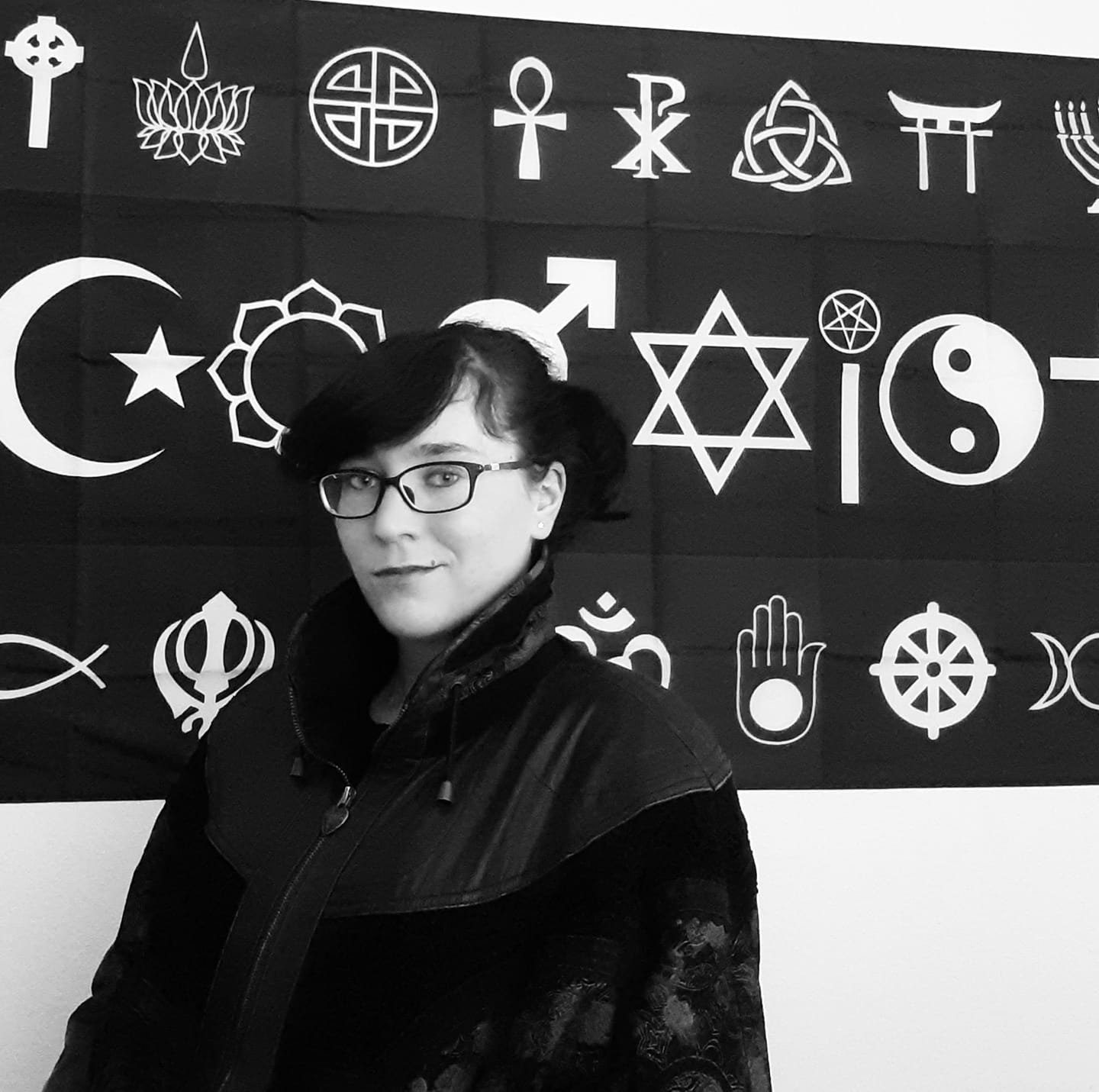 Person standing in front of a black banner displaying various religious and cultural symbols, including the crescent and star, Star of David, Dharma Wheel, and Yin Yang.