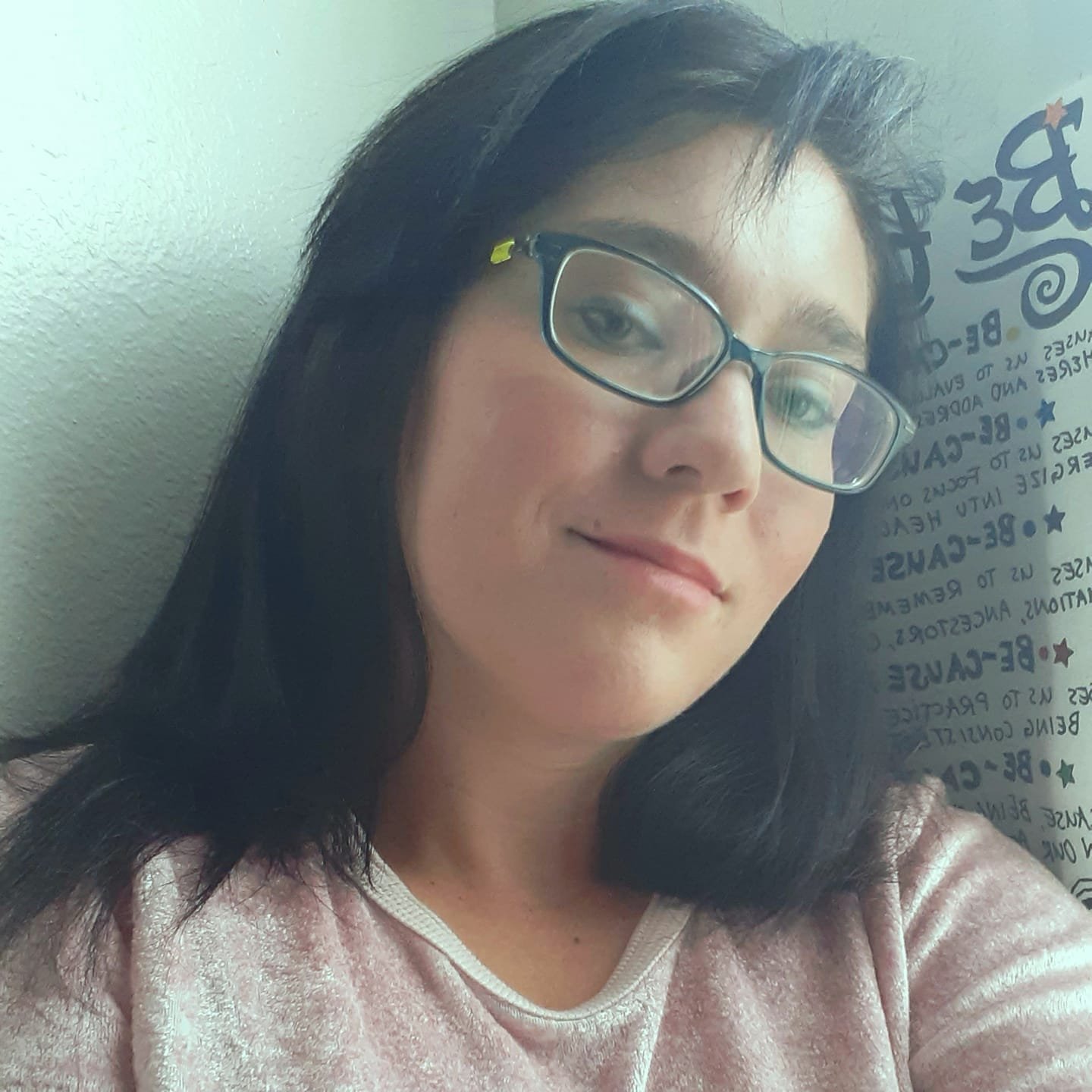 Person with glasses and dark hair smiling slightly, wearing a light-colored top, against a wall with text and decorative elements.
