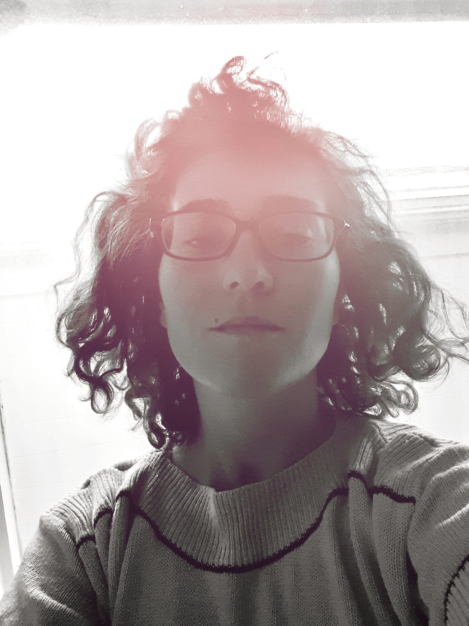 A person with curly hair and glasses in a black and white photo with a pink hue, wearing a sweater.