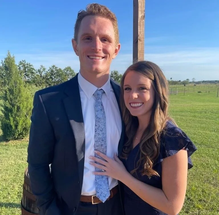 A young man in a suit and a young woman in a dress smiling outdoors on a sunny day, with green fields and trees in the background.