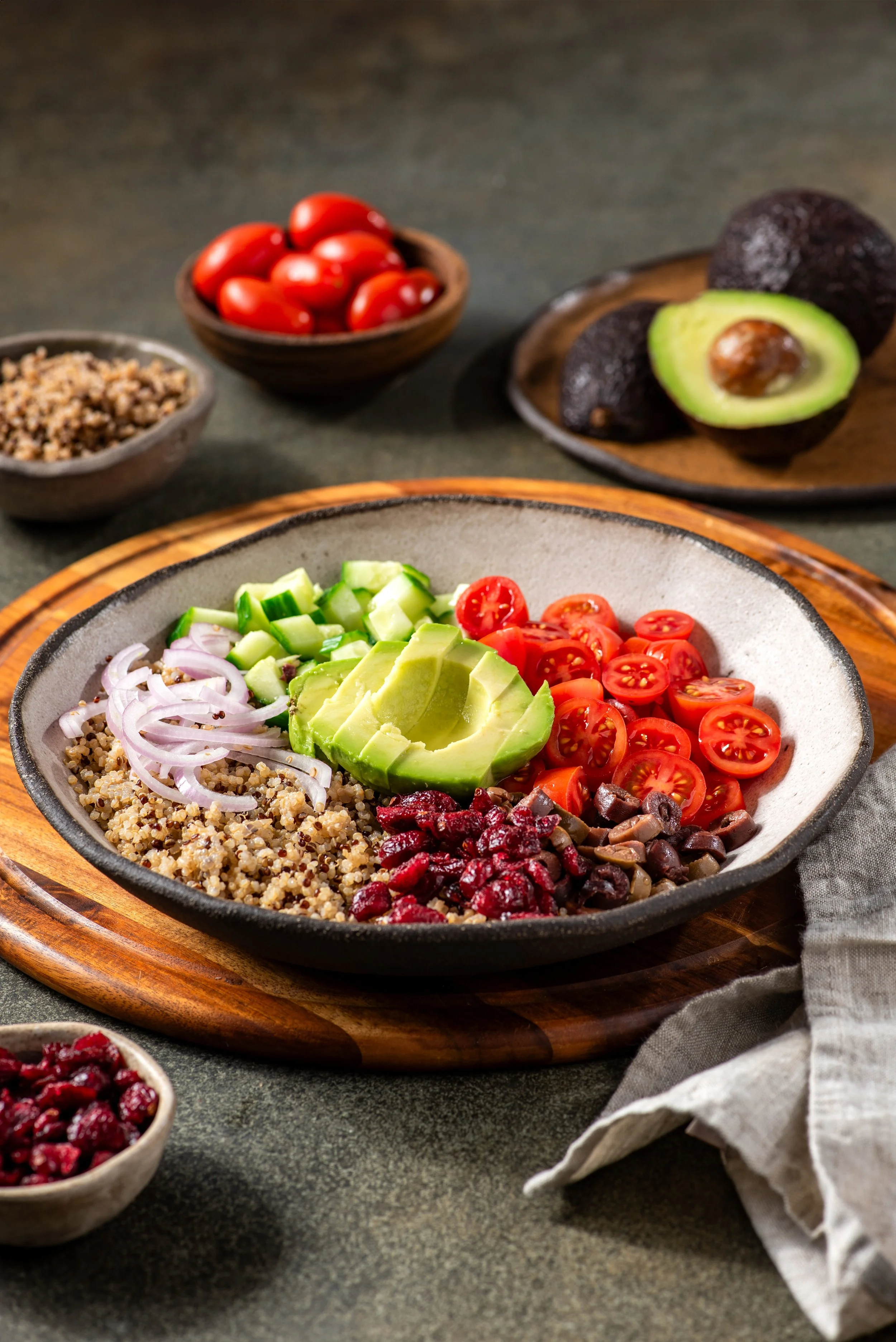 A bowl of quinoa salad topped with sliced avocado, cherry tomatoes, and red onions on a wooden platter, with bowls of cherry tomatoes and avocados in the background.