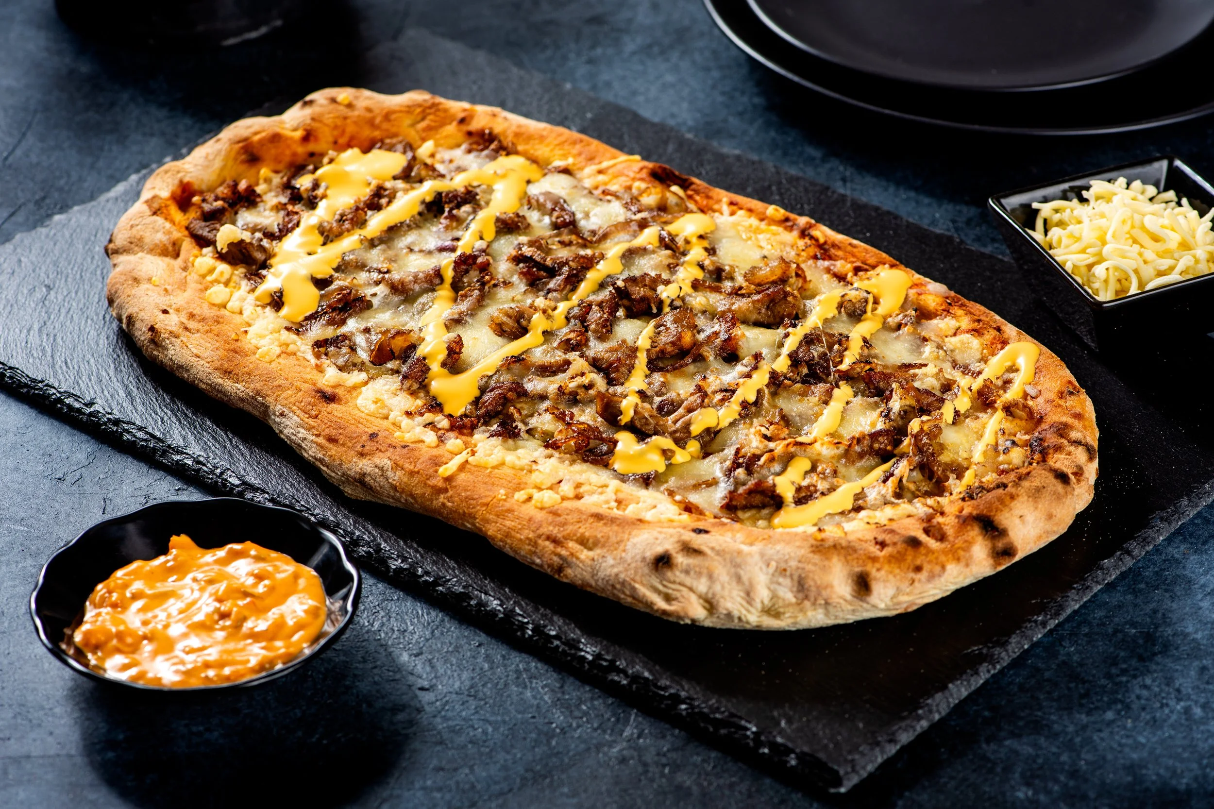 An elongated pizza with beef, cheese, and mustard on a dark surface, accompanied by a small bowl of cheese and a bowl of a creamy orange sauce.