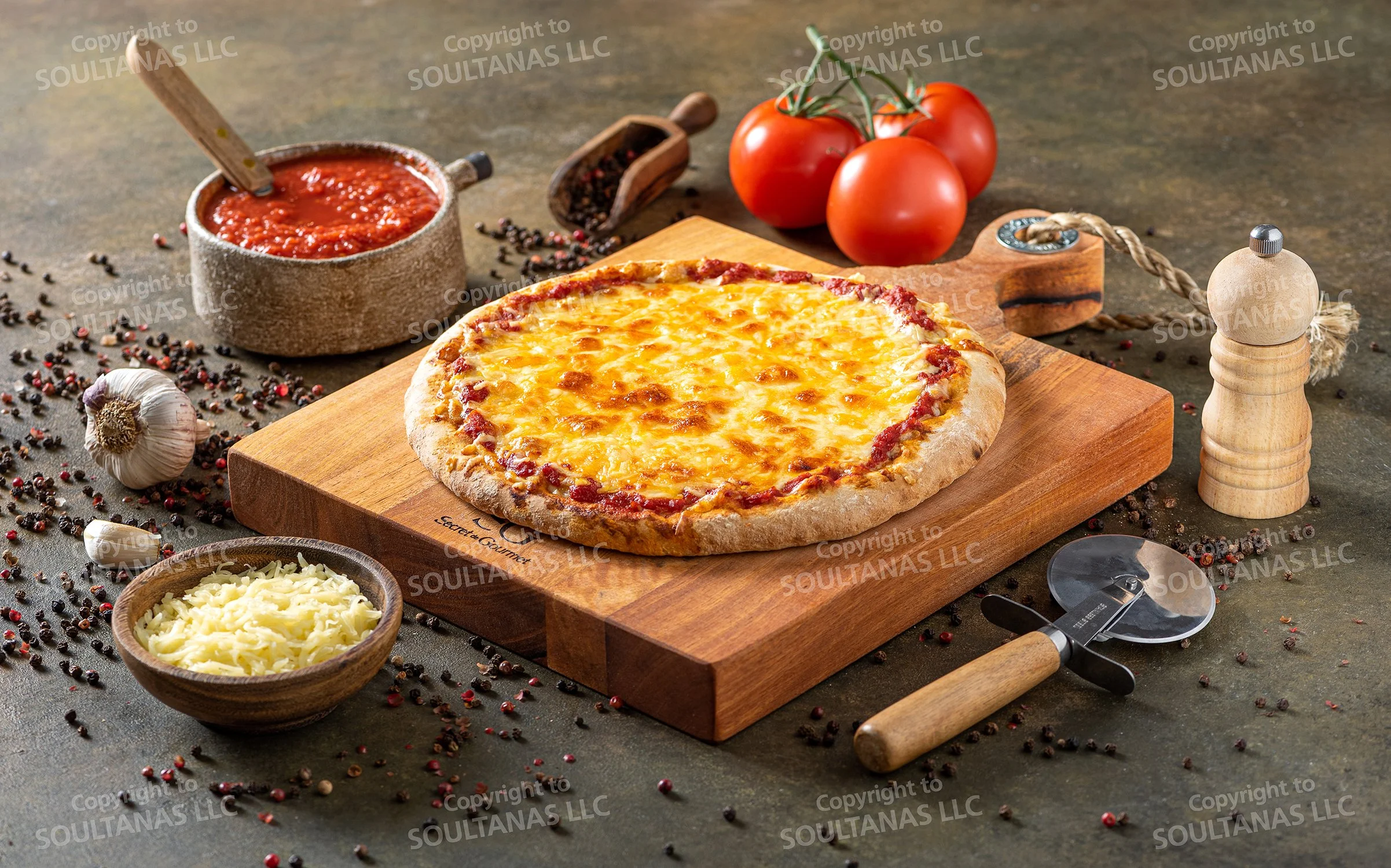 A freshly baked cheese pizza on a wooden board surrounded by tomatoes, garlic, shredded cheese, a bowl of tomato sauce, and a pizza cutter, with scattered peppercorns and a pepper mill in the background.