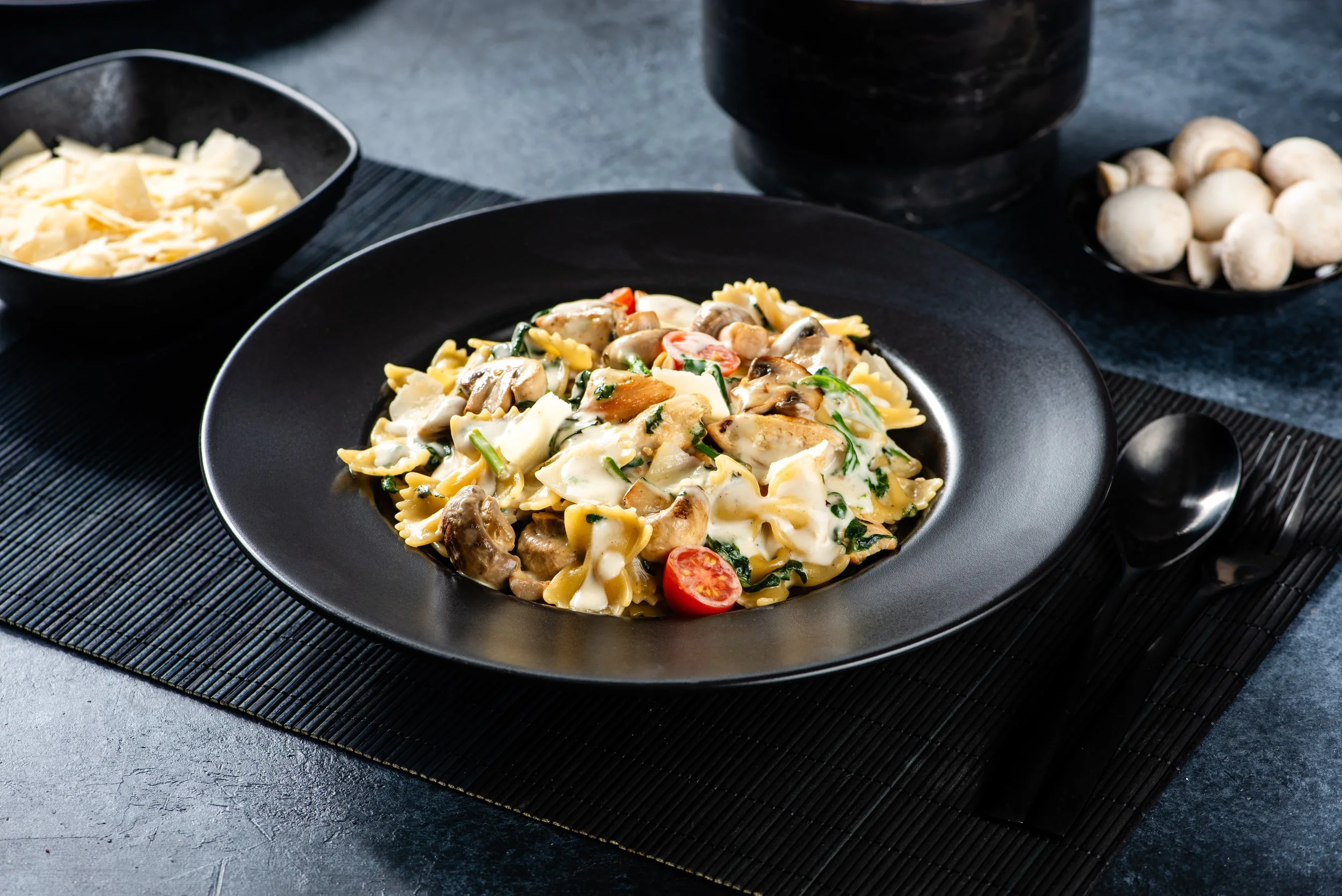 A black plate of creamy mushroom and cherry tomato pasta with spinach, served on a black placemat with bowls of shredded cheese and white button mushrooms in the background.
