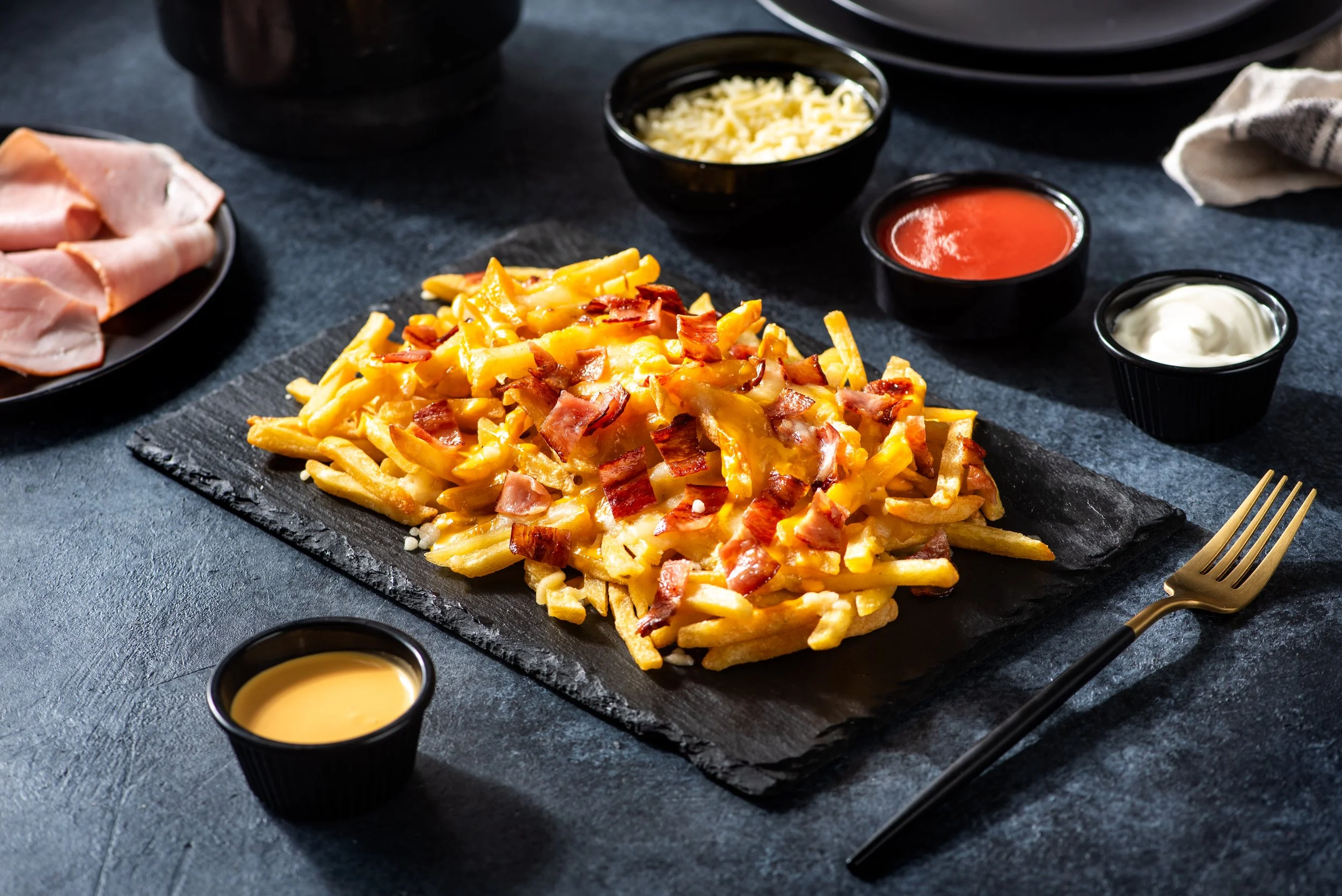 French fries topped with cheese and bacon, served on a black slate platter with small bowls of ketchup, mayonnaise, cheese sauce, and another creamy sauce on a dark surface.