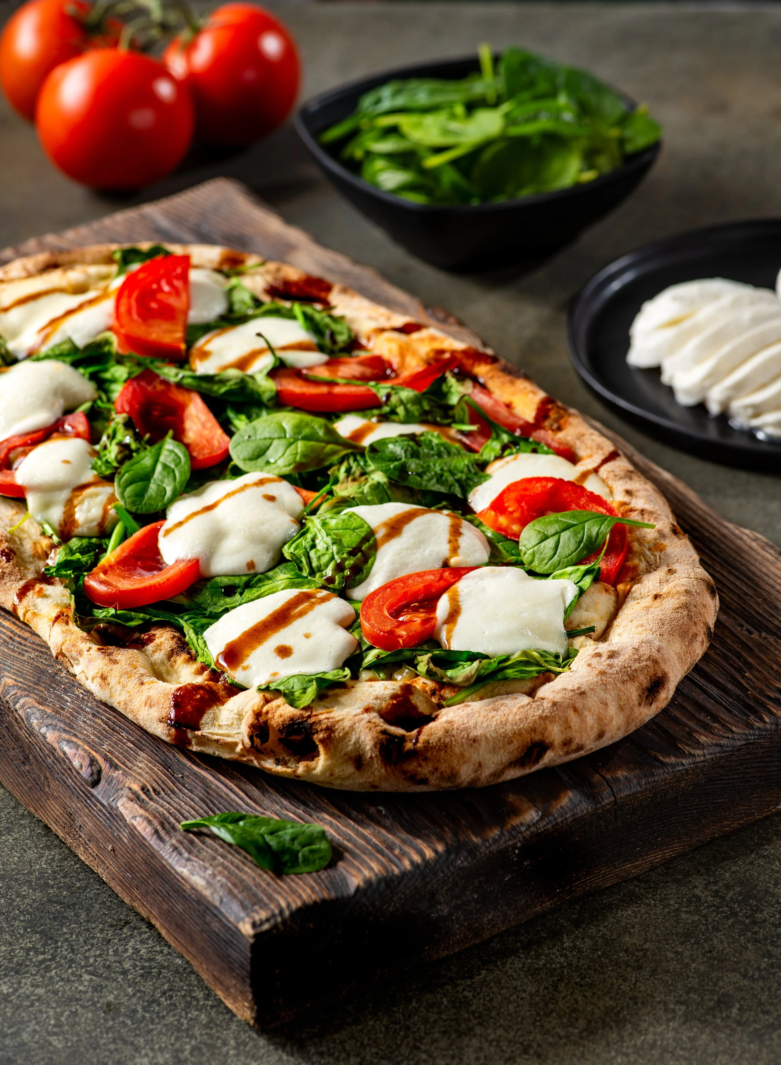 A close-up of a pizza topped with slices of tomato, mozzarella cheese, and fresh basil, sitting on a wooden board with cherry tomatoes and green leafy vegetables in the background.