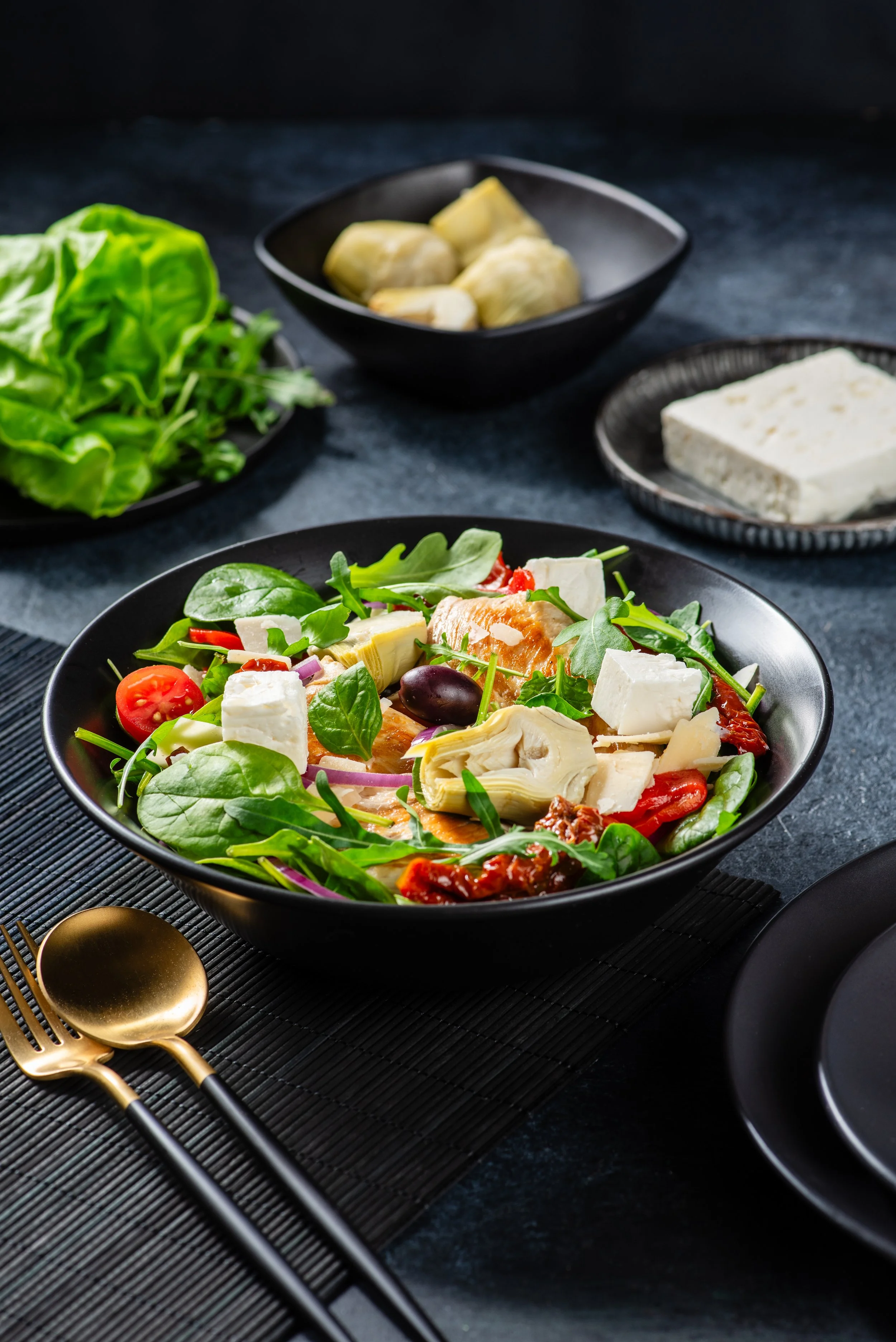 A bowl of fresh salad with cherry tomatoes, spinach, feta cheese, artichoke hearts, and sun-dried tomatoes, with side dishes of lettuce, artichoke hearts, and feta cheese on a dark textured surface.