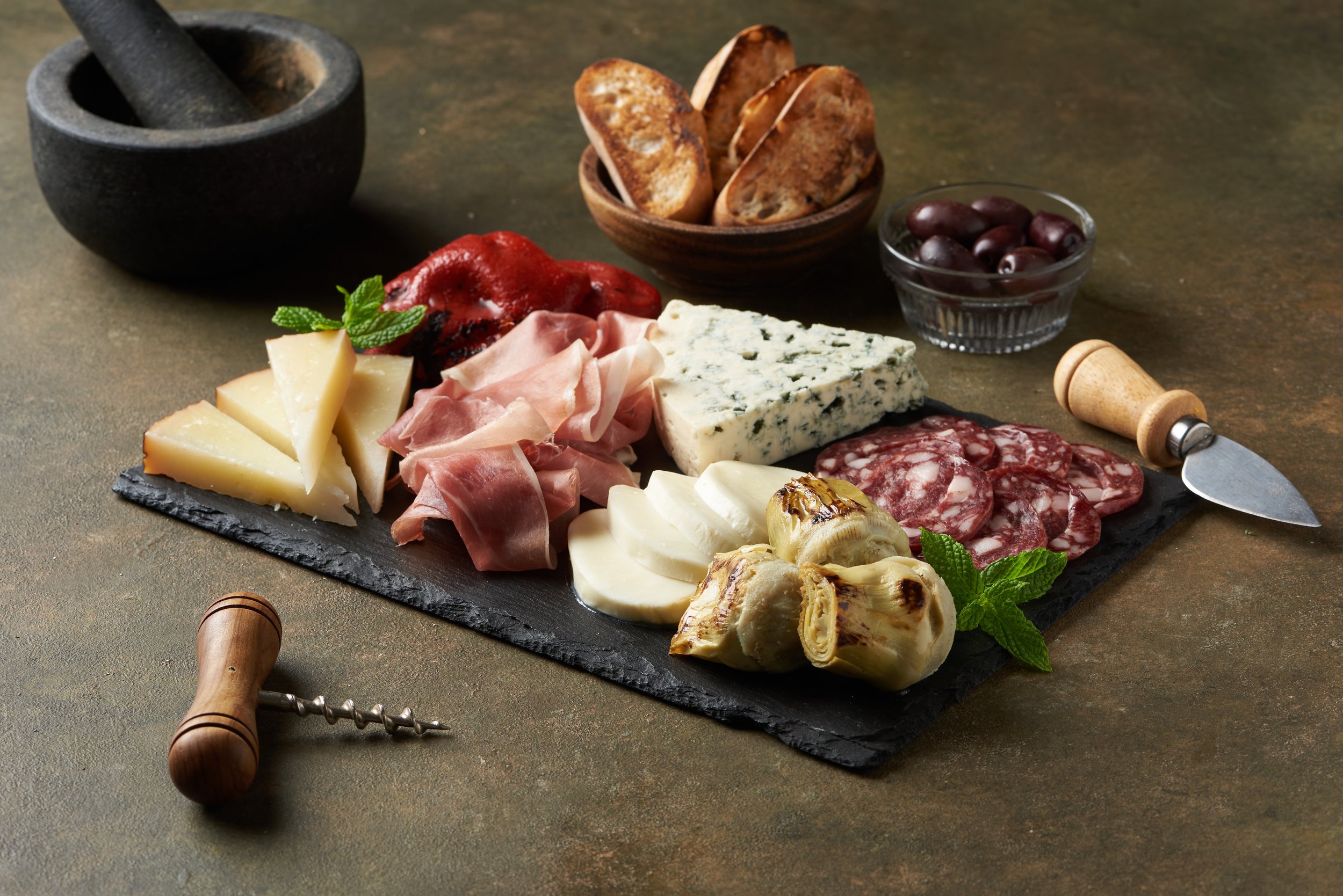 A variety of cheeses, cured meats, grapes, toasted bread slices, and garnishes arranged on a black slate serving board, with a mortar and pestle and a corkscrew nearby.