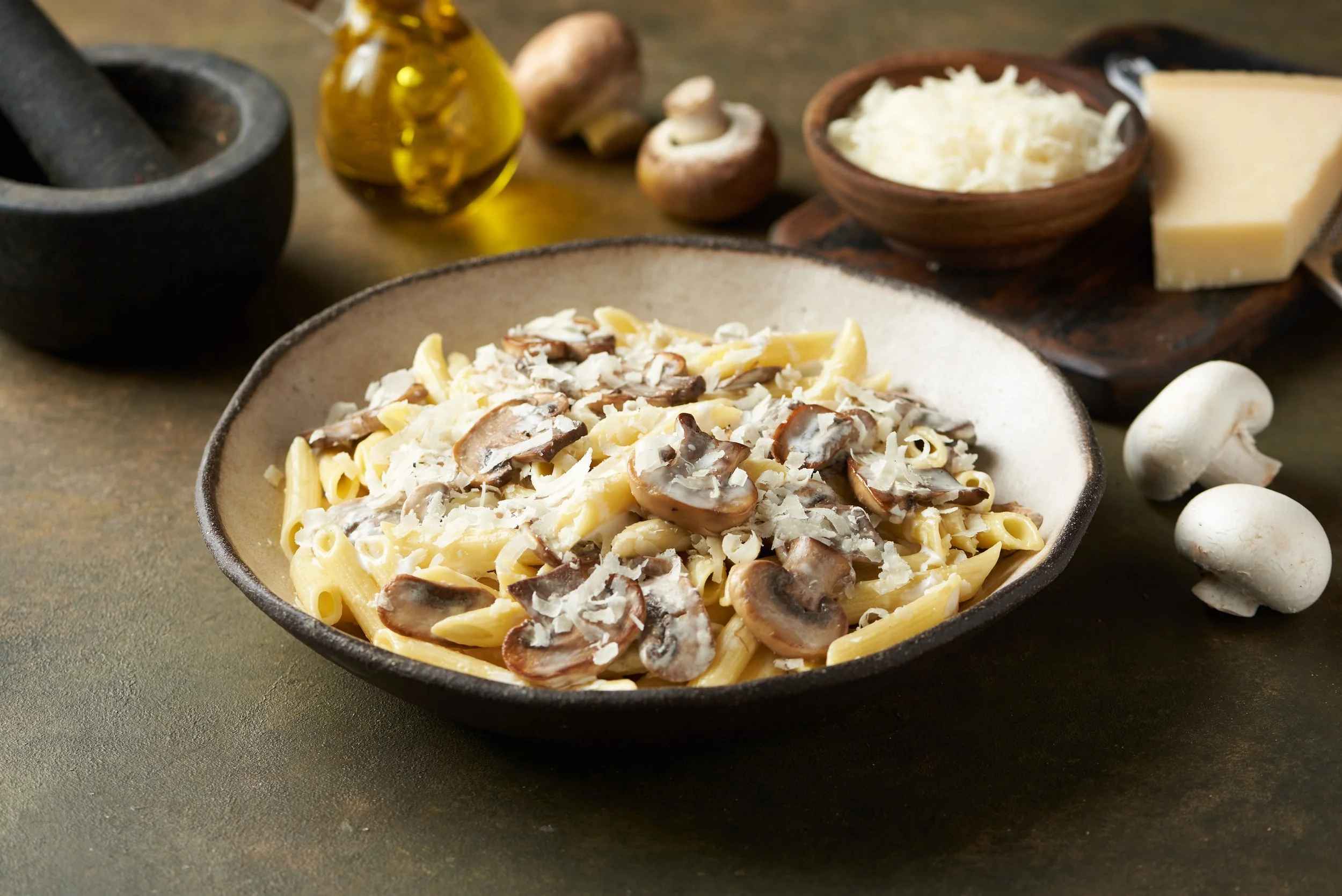 A bowl of cooked pasta with mushrooms and grated cheese, surrounded by fresh mushrooms, a small bowl of grated cheese, a bottle of olive oil, garlic cloves, and a mortar and pestle on a rustic surface.