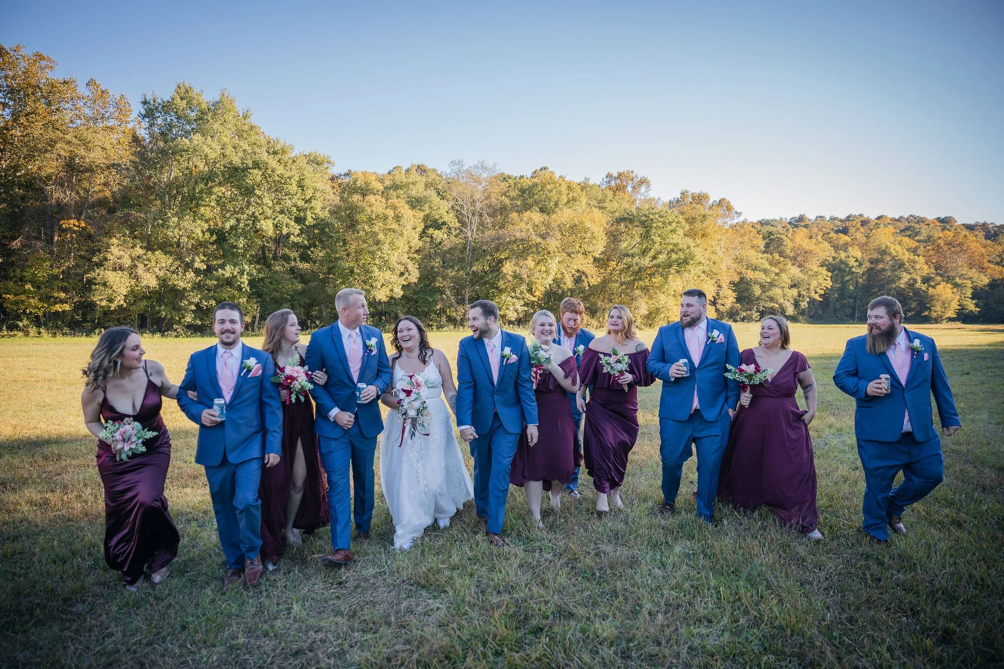A group of wedding guests walking together in a grassy field with trees in the background, dressed in formal attire, holding flowers and drinks, smiling and enjoying the outdoor event.