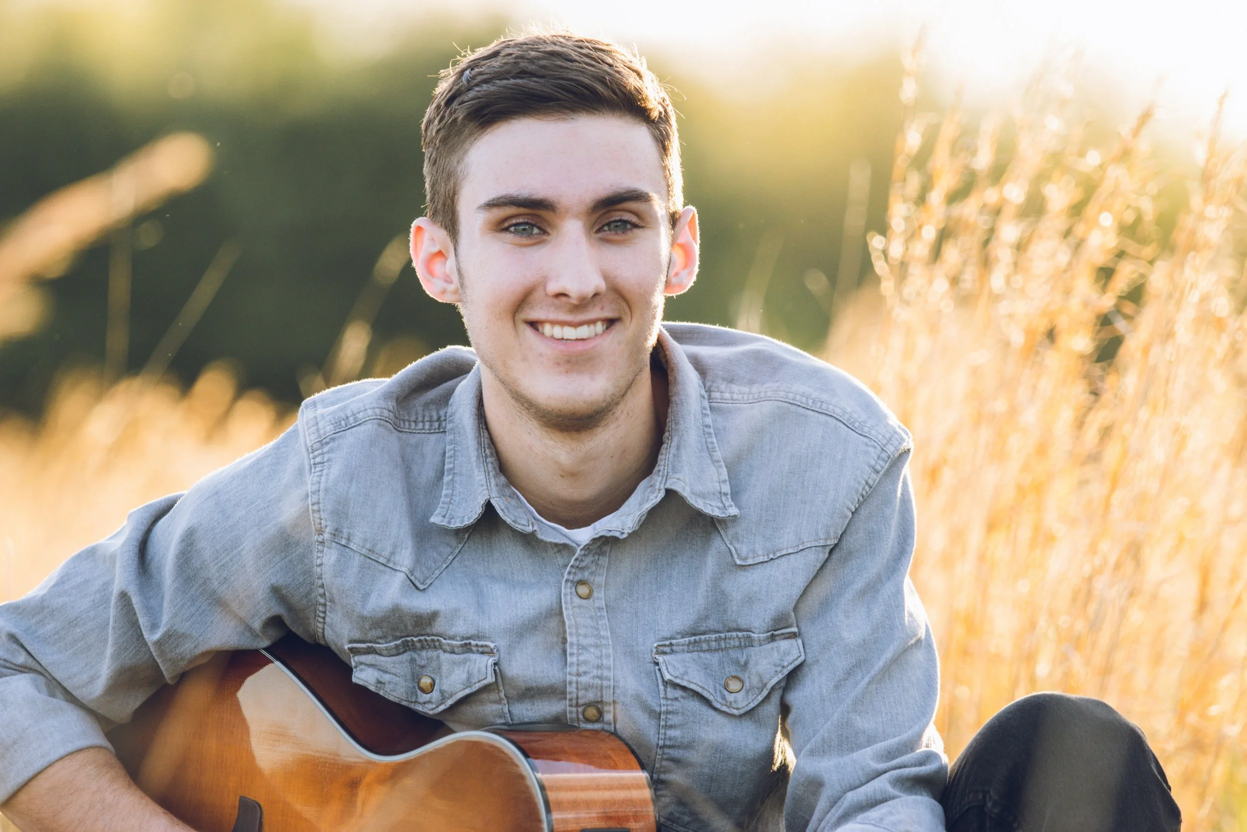 A young man smiling while sitting in a field of tall, golden grass with a guitar, during sunset.