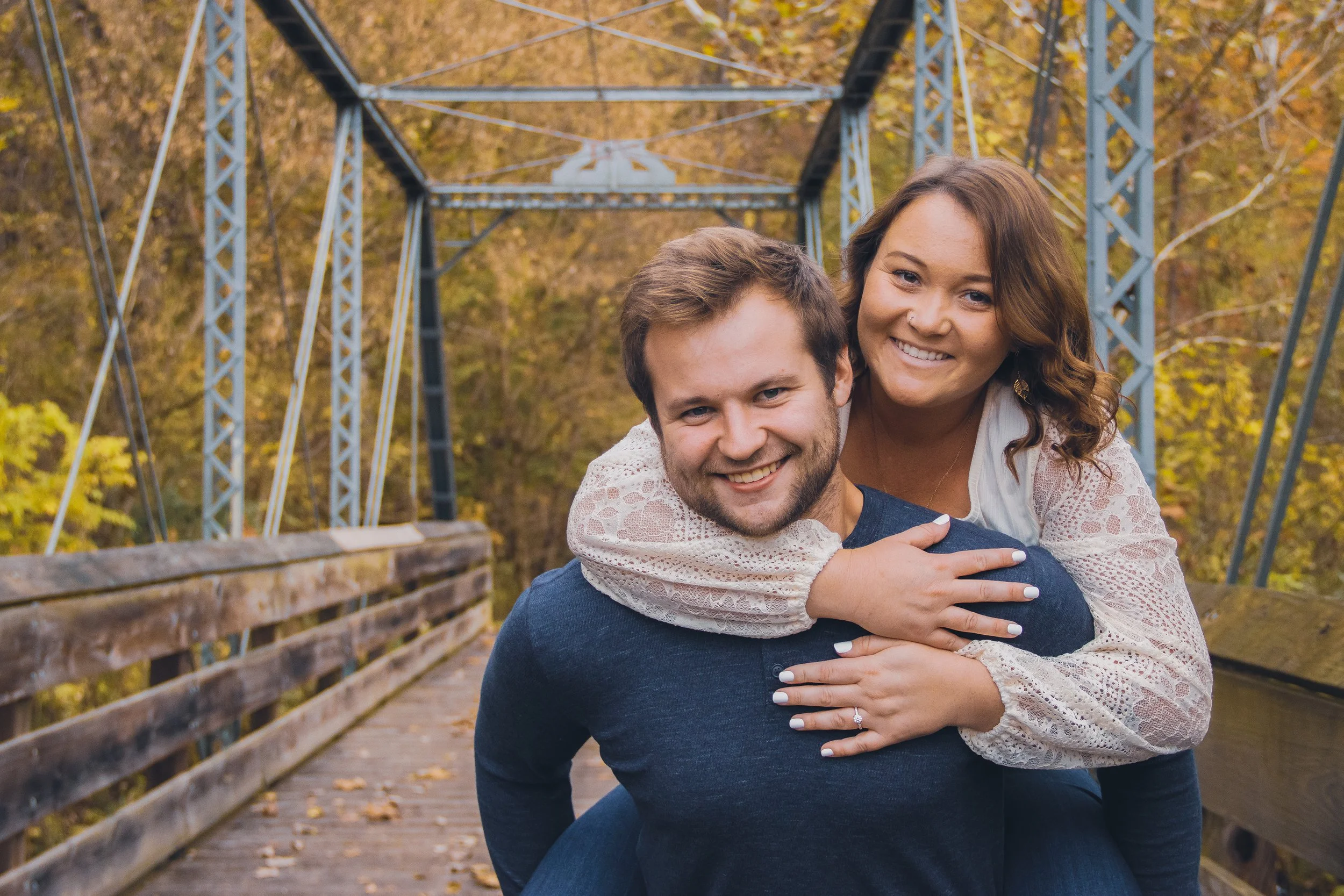 A smiling man giving a piggyback ride to a smiling woman on a wooden bridge during autumn.