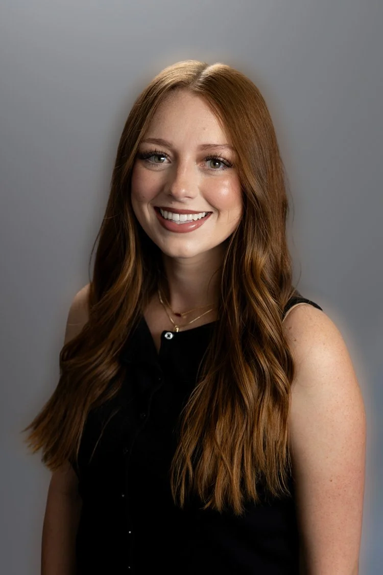 Young woman with shoulder-length red hair smiling outdoors, wearing a black t-shirt, layered necklaces, and a belt, with trees and grass in the background.
