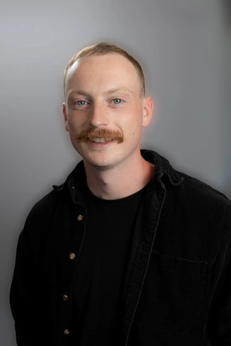 A young man with red hair, a mustache, and light skin, wearing a black T-shirt with a Vans logo and a brown cap, against a plain dark background.