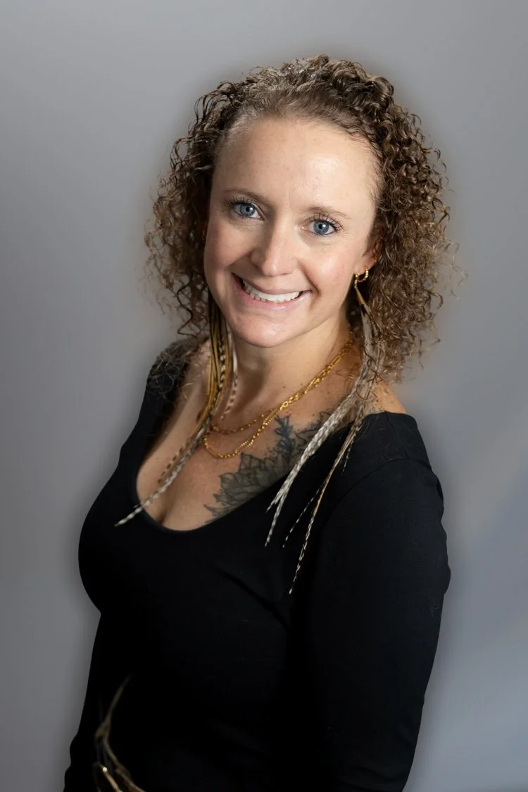 A woman with curly red hair, wearing a black lace top, smiling outdoors in a park or garden during golden hour.