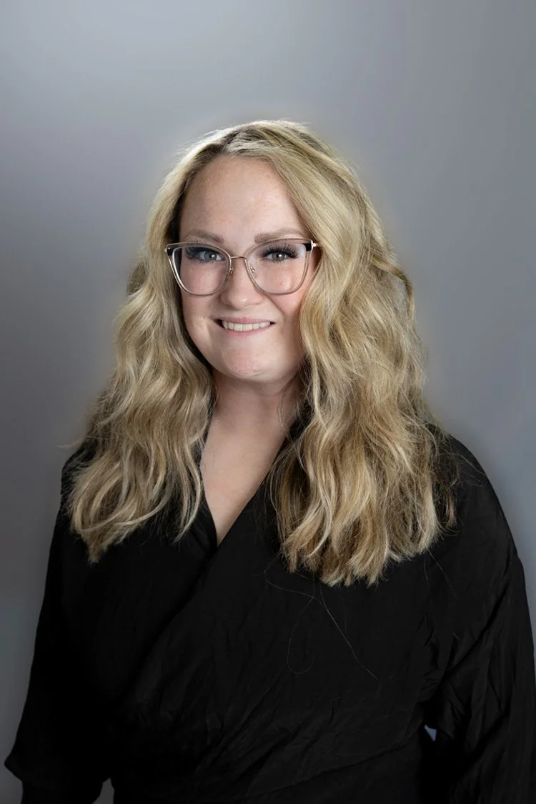 A woman with glasses smiling outdoors near a stream and rocky landscape.