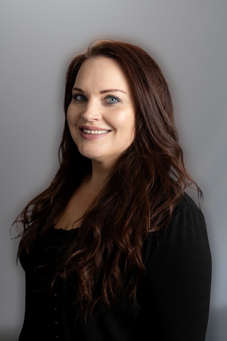 A woman with long, reddish-brown hair and blue eyes smiling outdoors during golden hour, wearing a black top with a zipper.