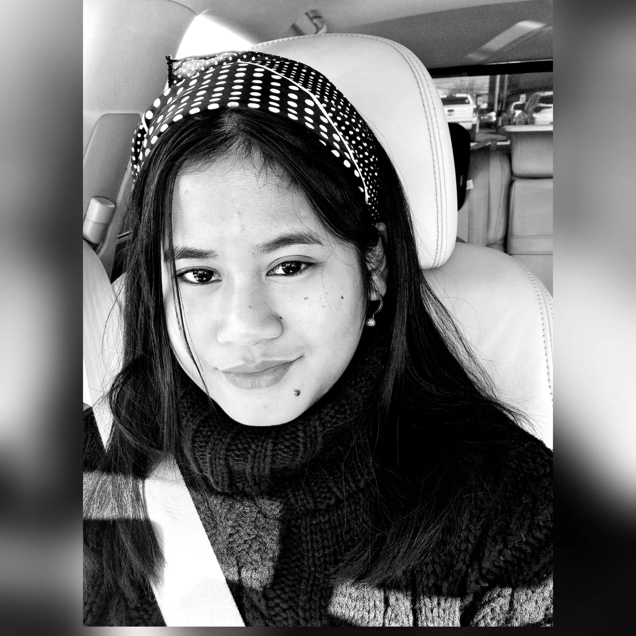 A woman with long dark hair wearing a patterned headband, earrings, and a knitted sweater, sitting in a car with a seatbelt on, taking a selfie.