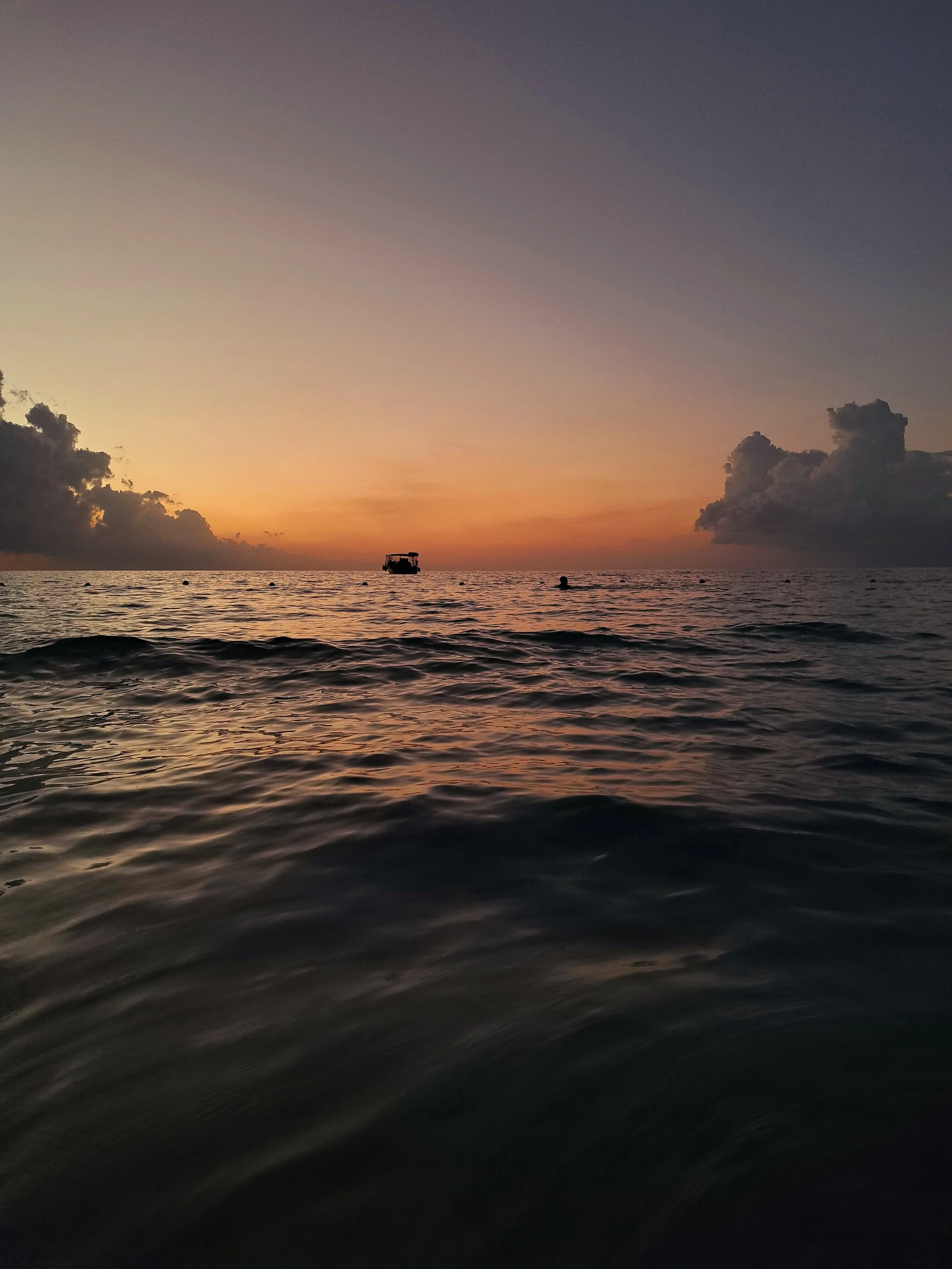 A photo of the ocean at sunset with a boat and a few people in the water, clouds in the sky, and a calm, rippling sea.