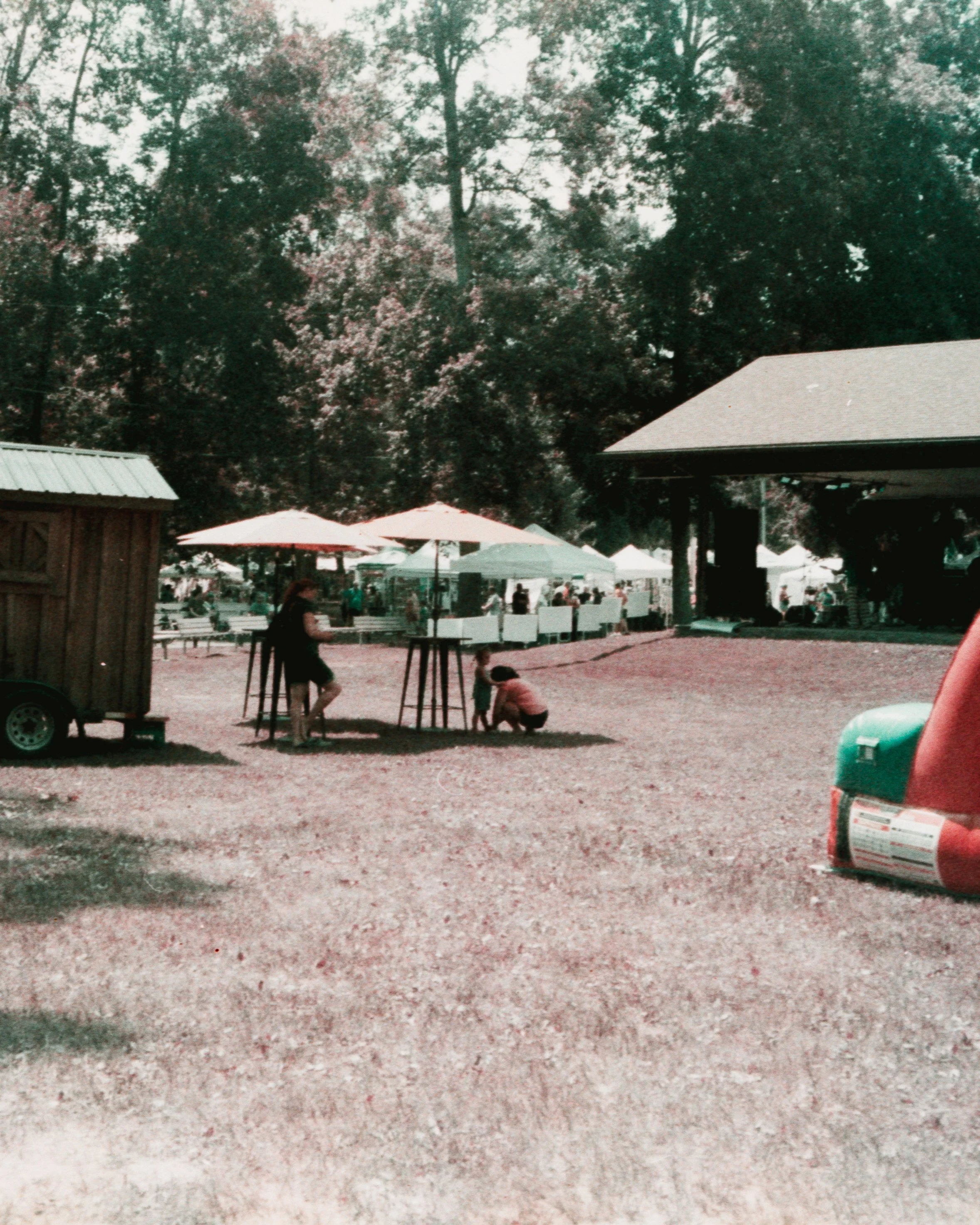 A daytime outdoor scene at a park or fair with trees, white tents, and people, including a child and a woman, near high tables and umbrellas.