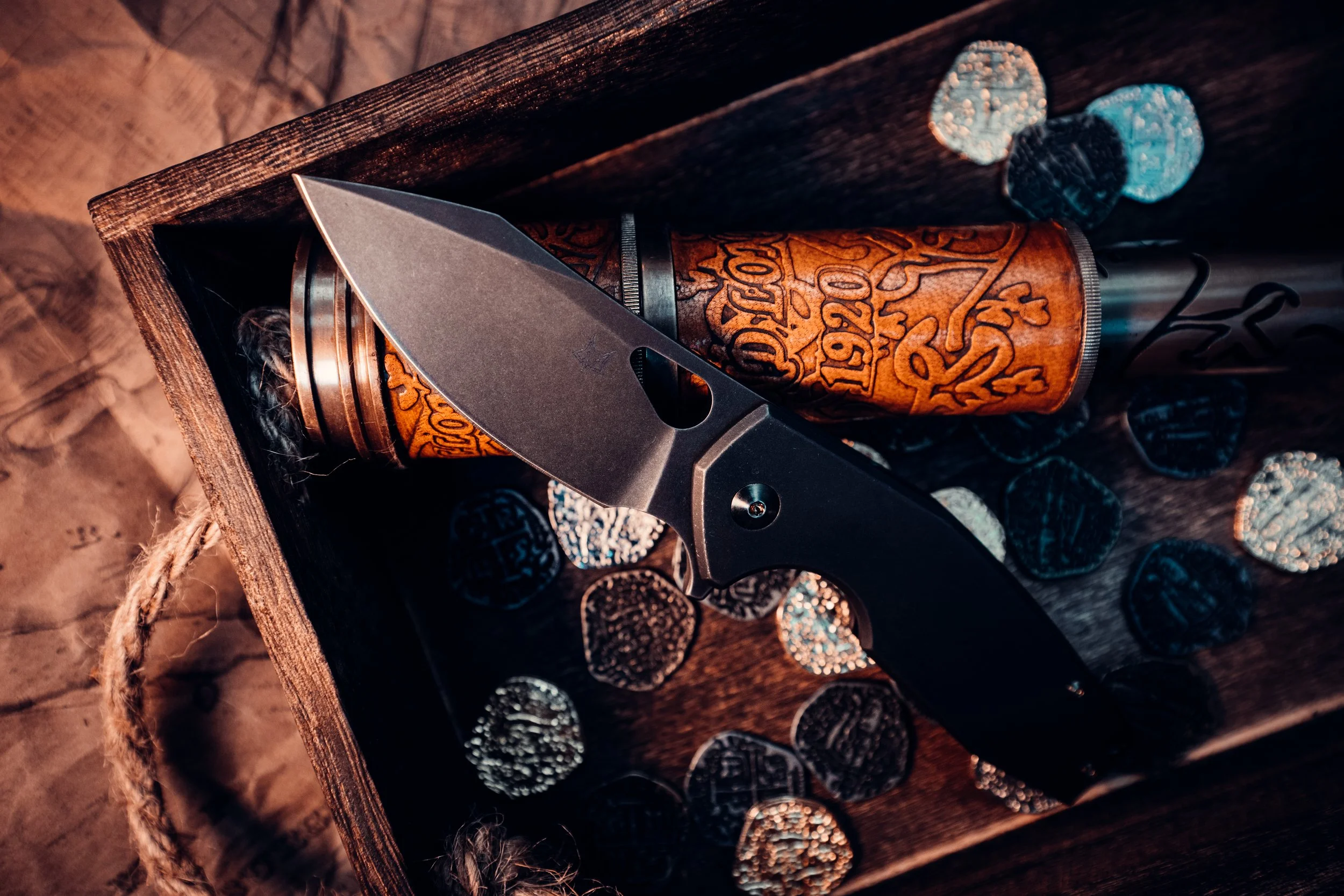 A black folding knife with a partially open blade placed inside a wooden box, which also contains a wooden object with carved designs and scattered silver coins.