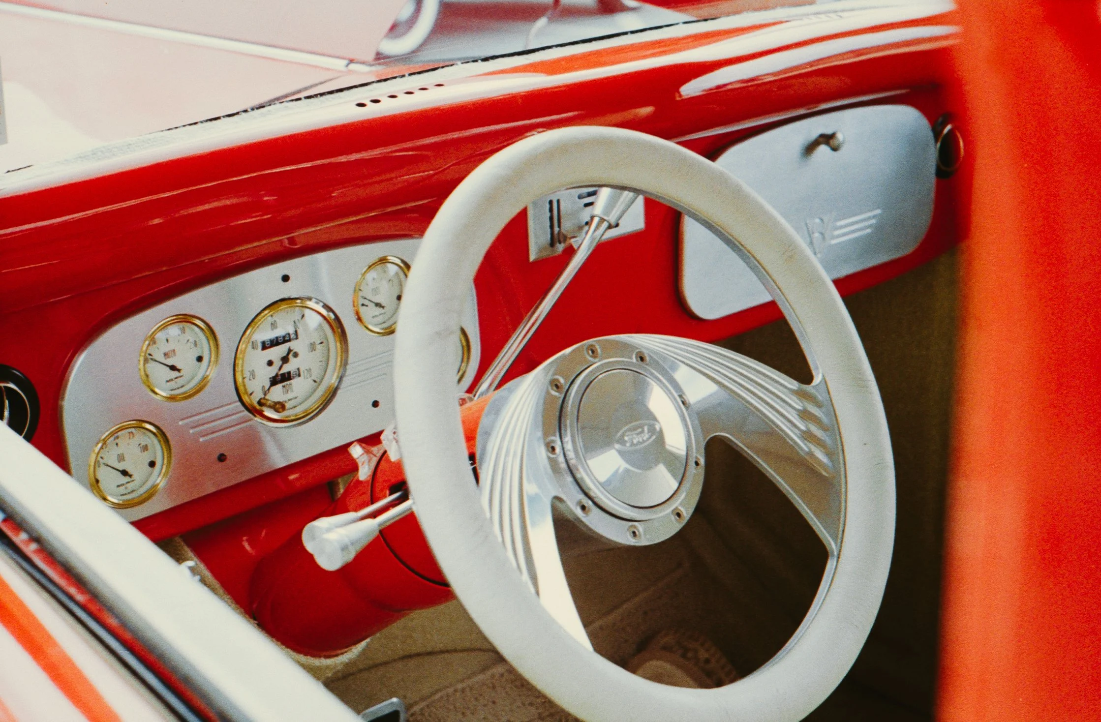 Close-up of a vintage car dashboard featuring white gauges with gold rims, a white steering wheel with metal spokes, and a red interior.