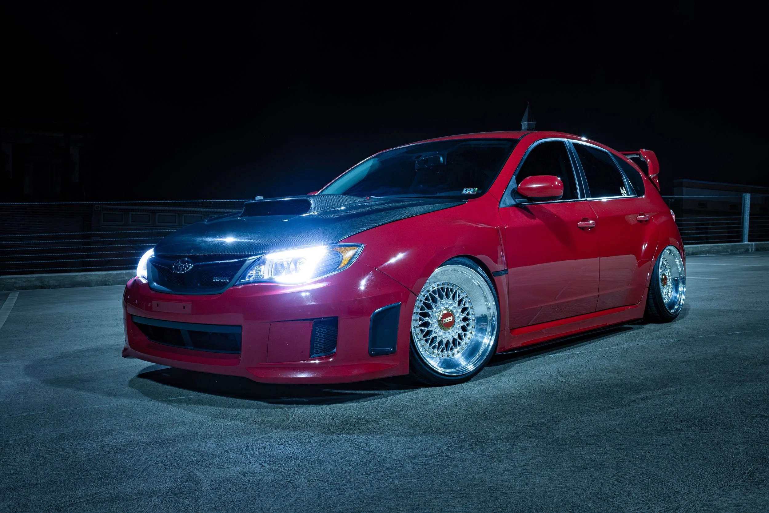A red modified car with black hood and shiny wheels parked on a rooftop at night.