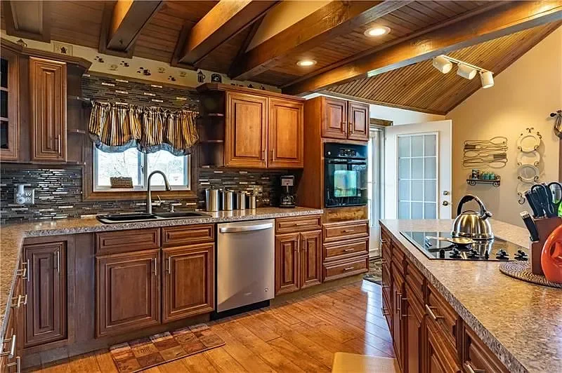 Real Estate Photography shot of a kitchen with wooden cabinets, a window with striped curtains, granite countertops, stainless steel appliances, and wooden ceiling beams.