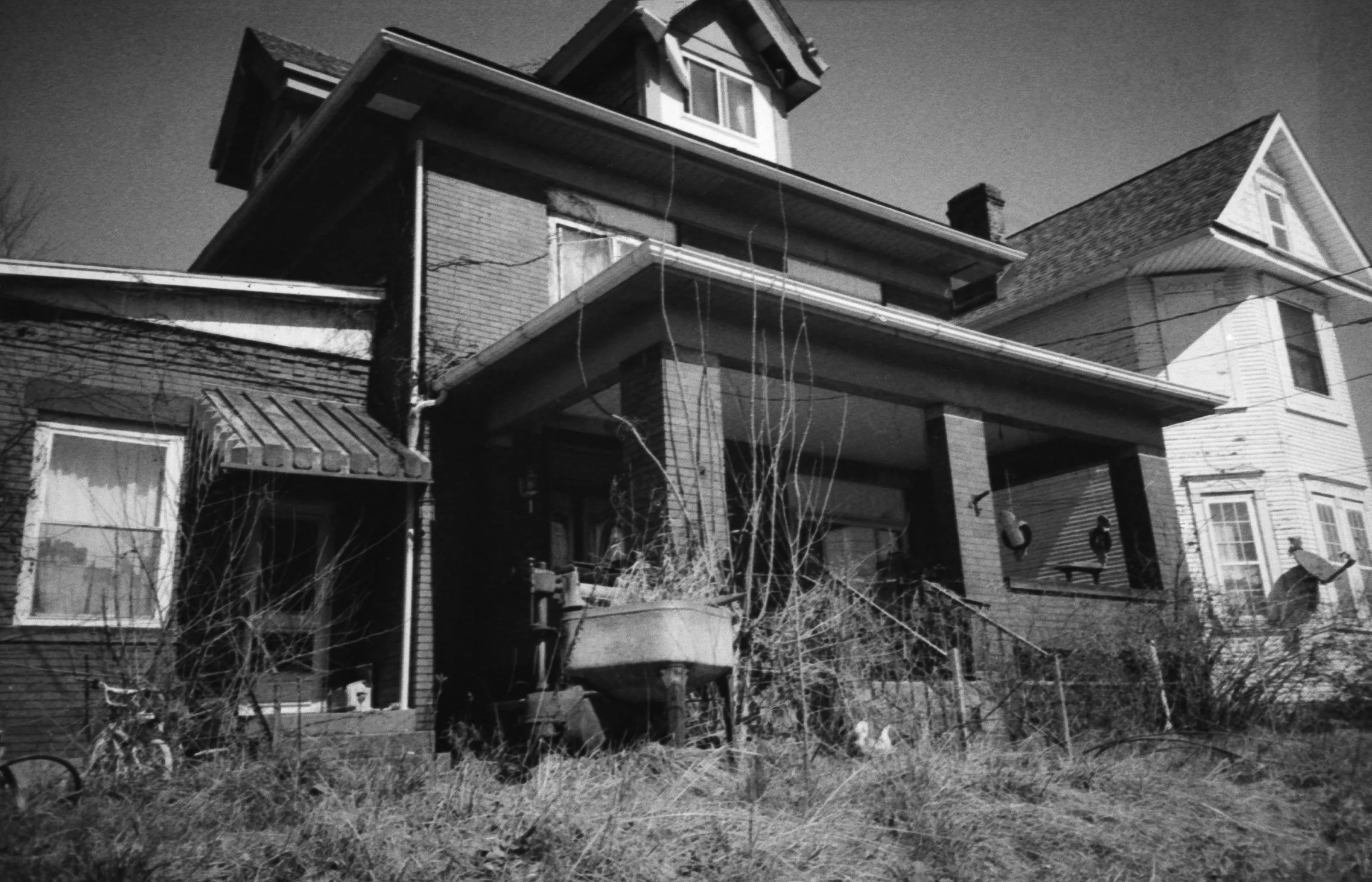 An old, weathered house with peeling paint and overgrown grass in the front yard, black and white photo.
